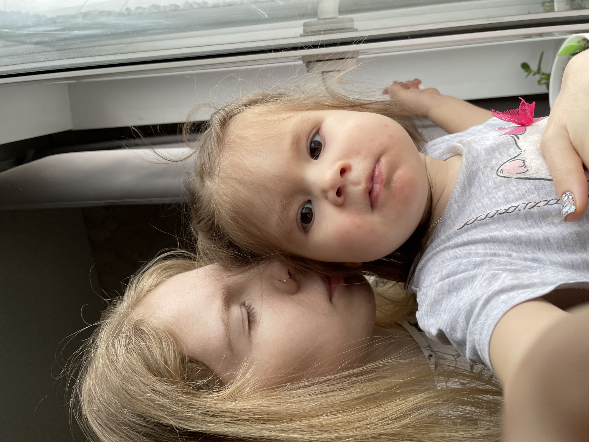 A selfie of a woman and a young child. The woman is lying on her back with blonde hair spread out. She has a nose ring and her eyes are closed. The child is lying on top of her, looking at the camera with light brown hair and a gray shirt with a pink bow. There are red spots on the child's face. The location appears to be indoors near a window. There is no information available to determine the location is in Izhevsk, Russia.