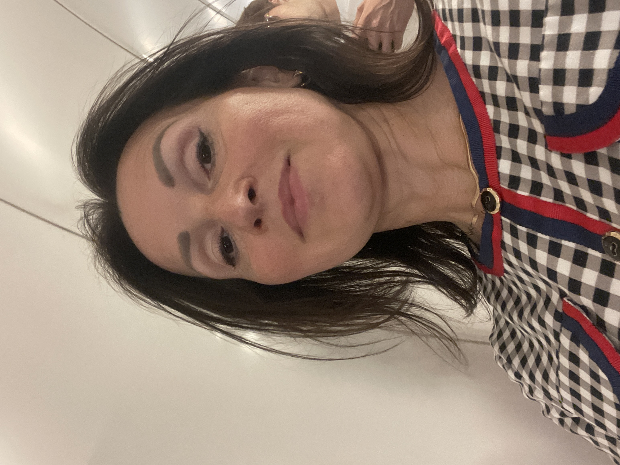 A woman with dark brown hair and fair skin takes a selfie. She is wearing a black and white checkered top with red and blue trim and gold buttons. She has on subtle makeup, including eyeliner and lipstick. Her hair is partially obscuring her face. A person's shoulder and arm are visible in the background to the right, with a hint of glasses. The background appears to be a white, possibly reflective surface, suggesting an indoor setting such as a hallway or room with mirrors. The lighting is bright, coming from above. The location is Unknown, Unknown.