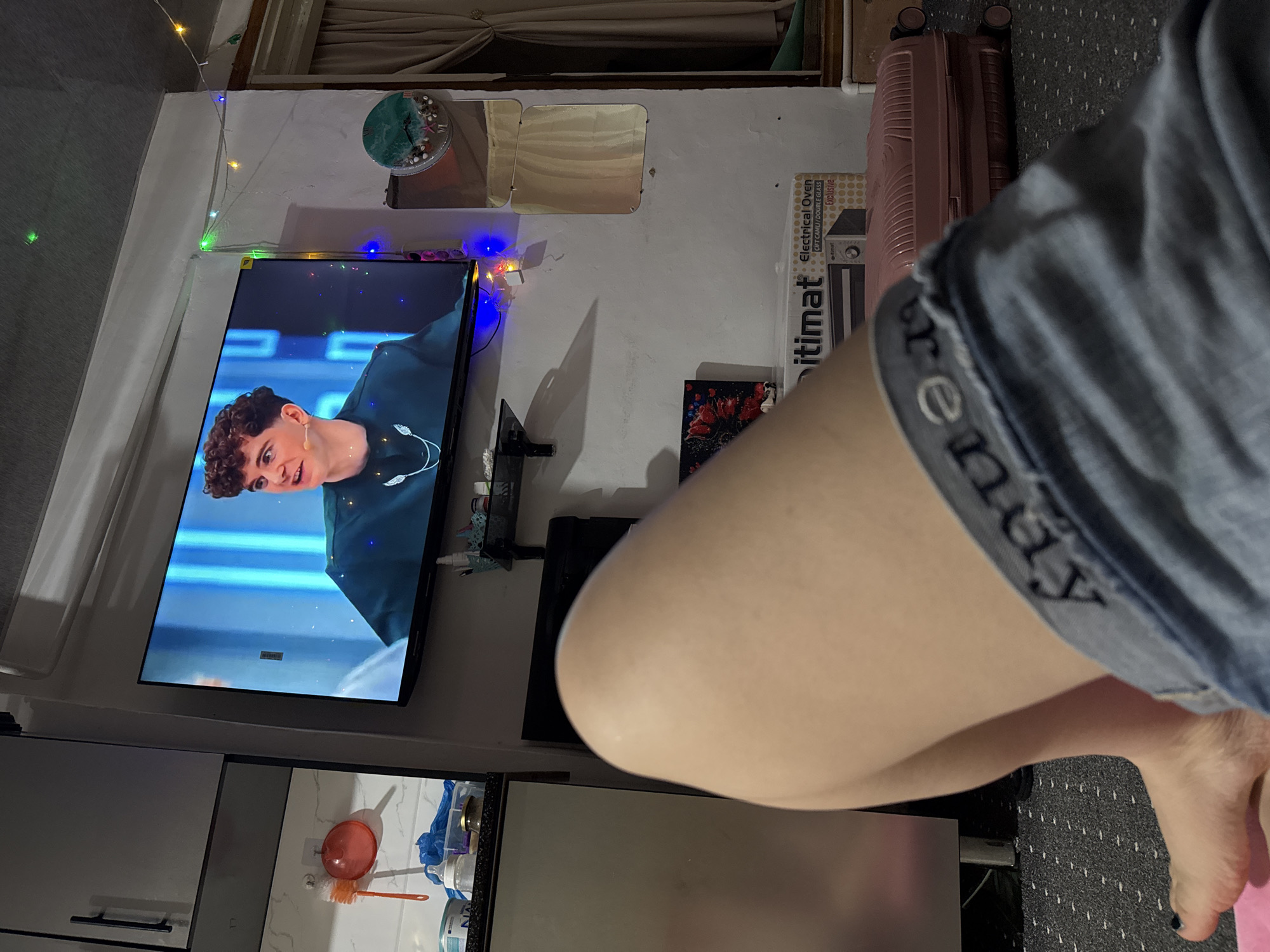In an indoor scene, a person's legs and feet, clad in denim shorts, are prominently in the foreground, suggesting a relaxed posture. The legs are smooth and pale, with one foot resting on a pink surface. Behind the legs, a television screen displays a young man with curly brown hair, wearing a dark green top with a necklace. The television is mounted on a wall that features Christmas lights. To the left of the television, a kitchen counter is visible with various items, including a bottle and a red brush. Further to the right, a pink suitcase and a boxed oven are present. The room appears to be a living space or bedroom, with a carpeted floor. The location is identified as Chardara, Uzbekistan.