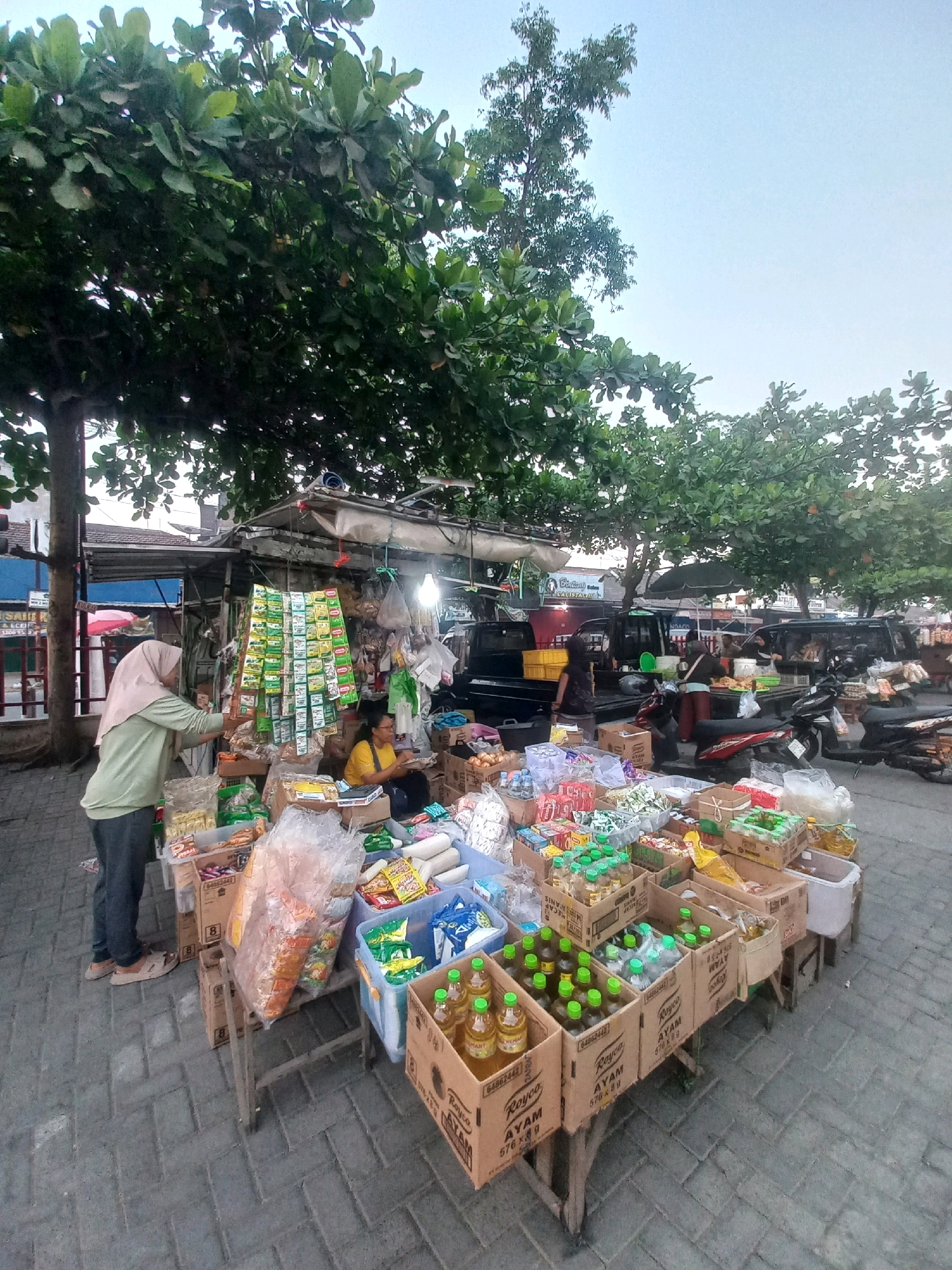 The image shows an outdoor market stall in Baturetno, Indonesia, likely during the day. A woman wearing a light green long-sleeved shirt and a beige hijab is standing on the left, examining products displayed on a rack. She is wearing dark pants and light brown shoes. In the center of the image, another woman with dark hair and a yellow shirt is seated behind the stall, appearing to be attending to customers or organizing goods.
The stall is laden with various items, including boxes of packaged food, bottles of drinks with green caps, and bags of snacks. Some items are displayed on wooden tables and shelves, while others hang from the stall's structure. Behind the stall, a dark-colored truck and several motorcycles are visible, suggesting a bustling market environment. The ground is paved with interlocking bricks. Trees with dense green foliage partially shade the scene, and the sky is overcast.