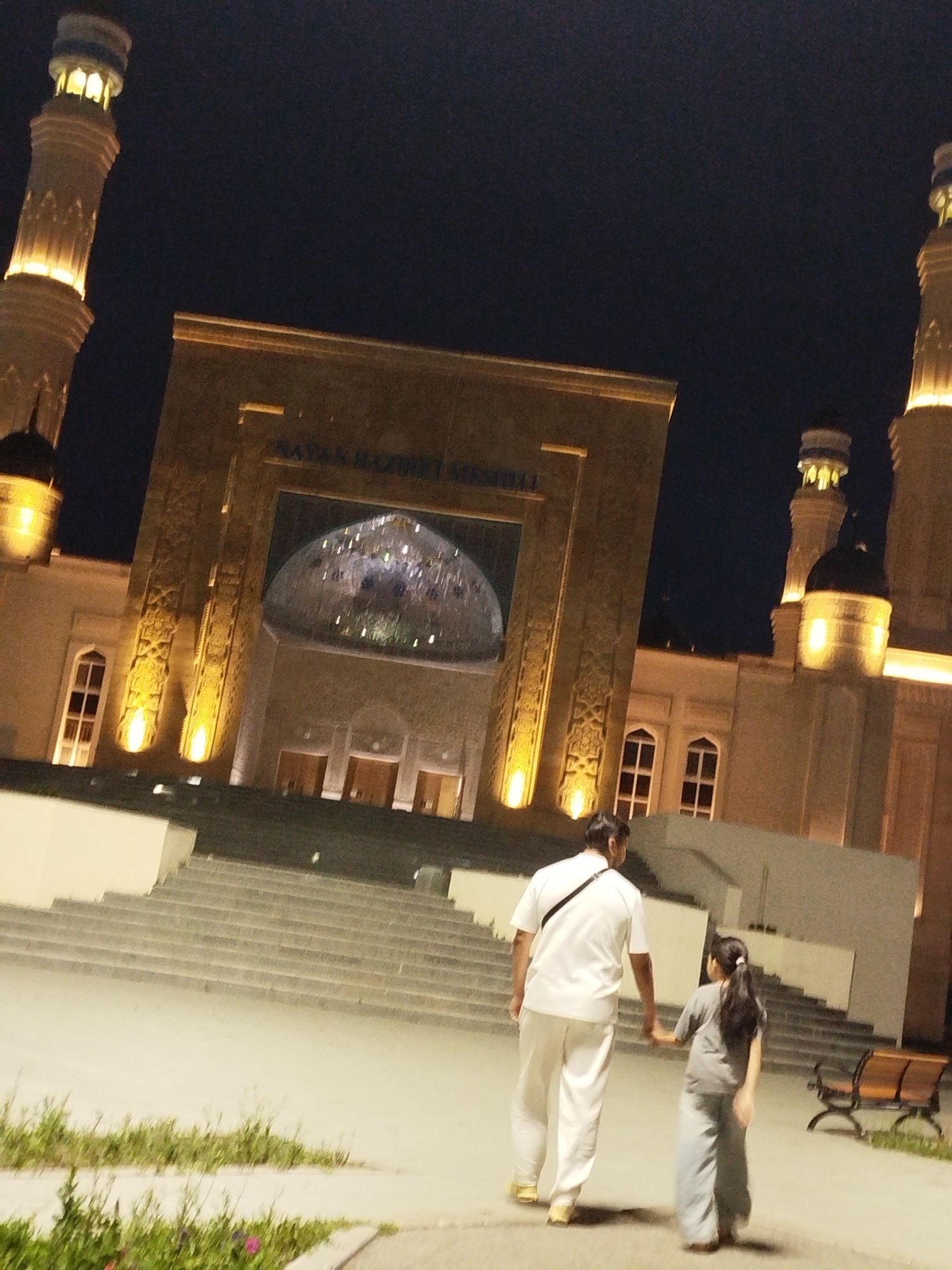 The image captures a nighttime view of the Nur-Astana Mosque in Kokshetau, Kazakhstan. The mosque's illuminated minarets and entrance are visible against a dark sky. In the foreground, a man and a young girl, presumably father and daughter, are walking away from the camera, holding hands. They are dressed in casual attire. The man is wearing a white shirt and pants, while the girl is in a grey shirt and pants. A park bench is visible in the background. The scene suggests a peaceful evening walk near a prominent religious landmark.