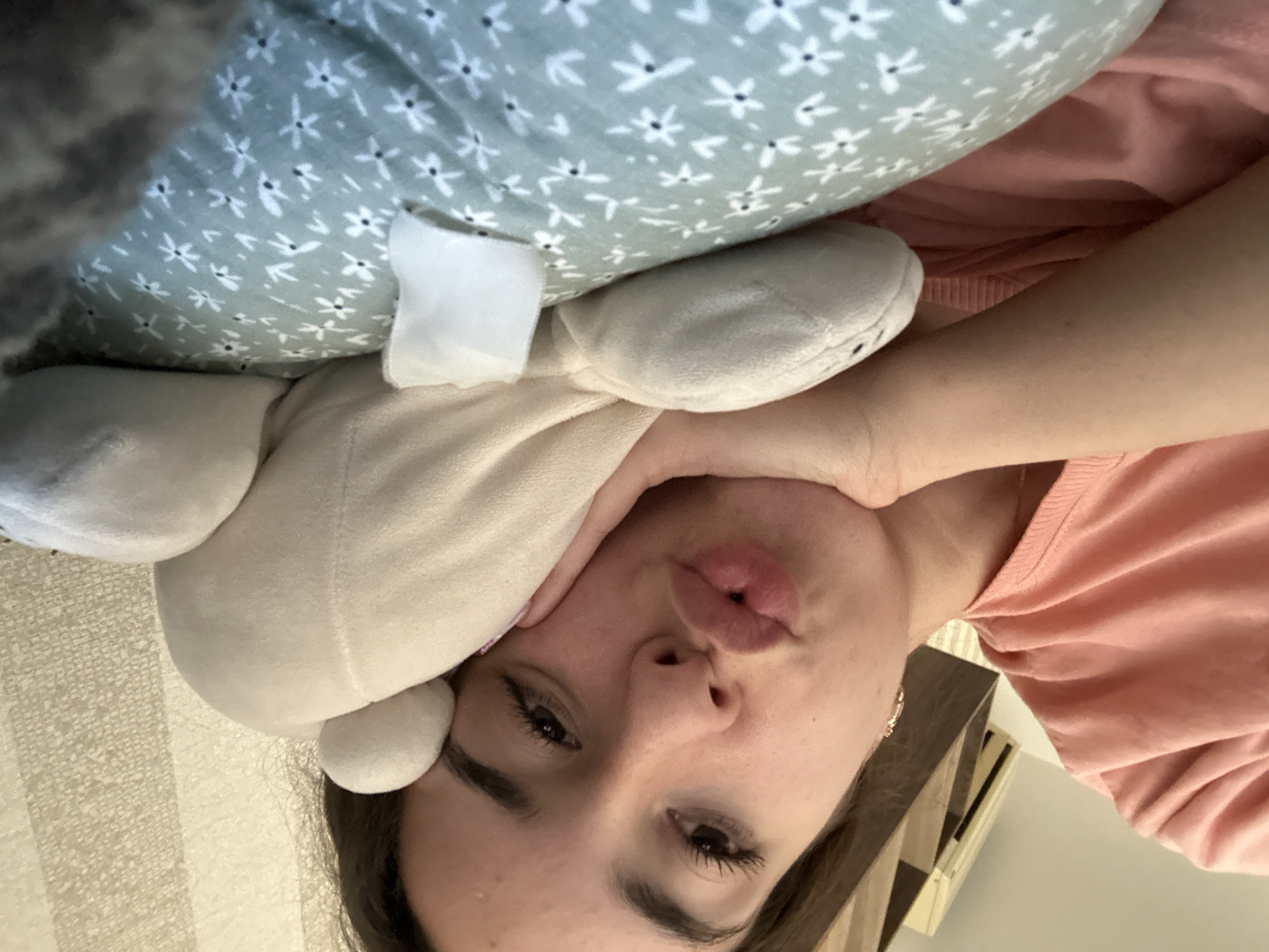 A young woman is lying on her side, propped up by a pillow. She is wearing a pink t-shirt and has her hand resting on a stuffed animal. Her lips are puckered, as if she is about to kiss the stuffed animal. The background is a textured wall, and there is a piece of furniture in the upper left corner. The image appears to be taken indoors, in a bedroom or living room. There are no distinctive landmarks that would indicate the specific location in Shumen, Bulgaria.