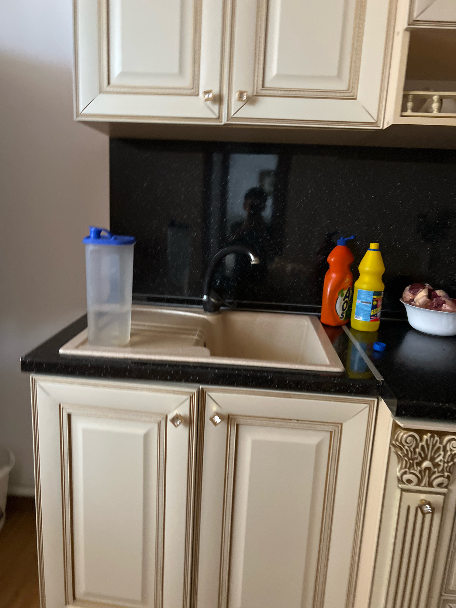 In Novogagatli, Russia, a kitchen sink area is depicted. A clear plastic water bottle with a blue lid, filled with water, stands on the left side of the sink. The sink itself is a light beige color and is positioned below a black, speckled countertop. To the right of the sink, two cleaning product bottles are visible: an orange bottle with "AOS" branding and a yellow bottle. Further to the right, a white bowl contains raw meat. Above the countertop, cream-colored cabinets with gold trim are installed. The background features a dark, reflective surface, possibly a backsplash, and the faint reflection of a person is visible. The lower portion of the image shows the front of cream-colored kitchen cabinets with decorative gold trim and diamond-shaped handles.