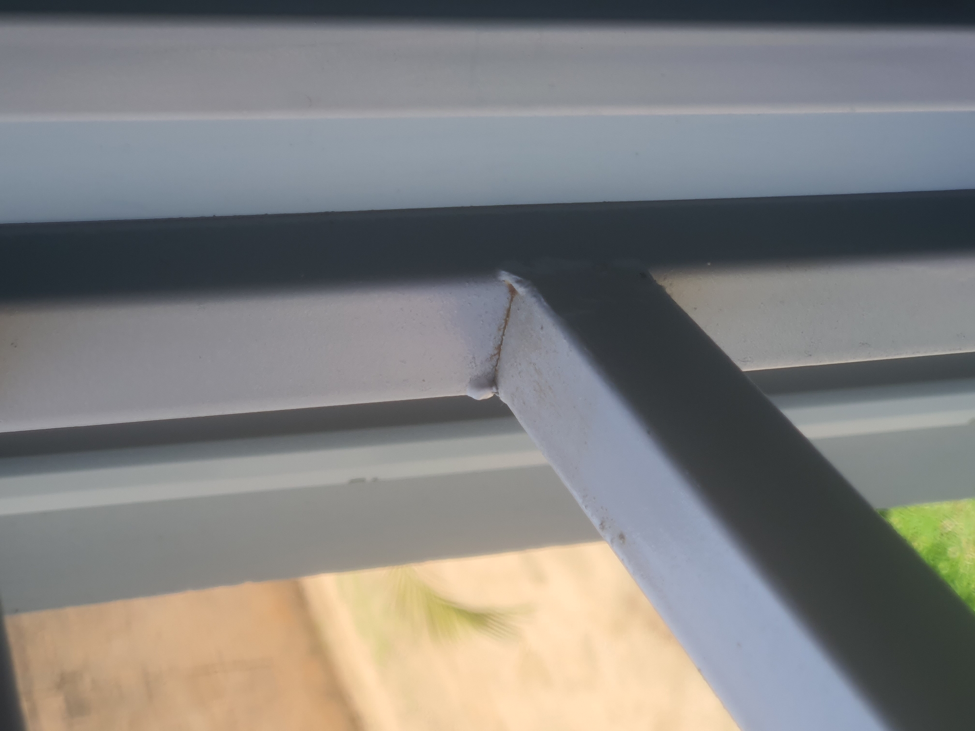 The image is a close-up shot of a light grey metal window grill. The grill consists of horizontal and vertical bars, with a visible weld joint in the foreground. The bars appear to be made of hollow metal tubing. Behind the grill, through the window, a blurred outdoor scene is visible. This includes what appears to be a sandy or dusty ground, some green grass, and a palm frond. The background wall is a light tan color. The overall impression is that of a residential or institutional building, likely in a warm climate. The image quality is moderate, with some focus blur, especially in the background. There are no people or animals visible in the frame.