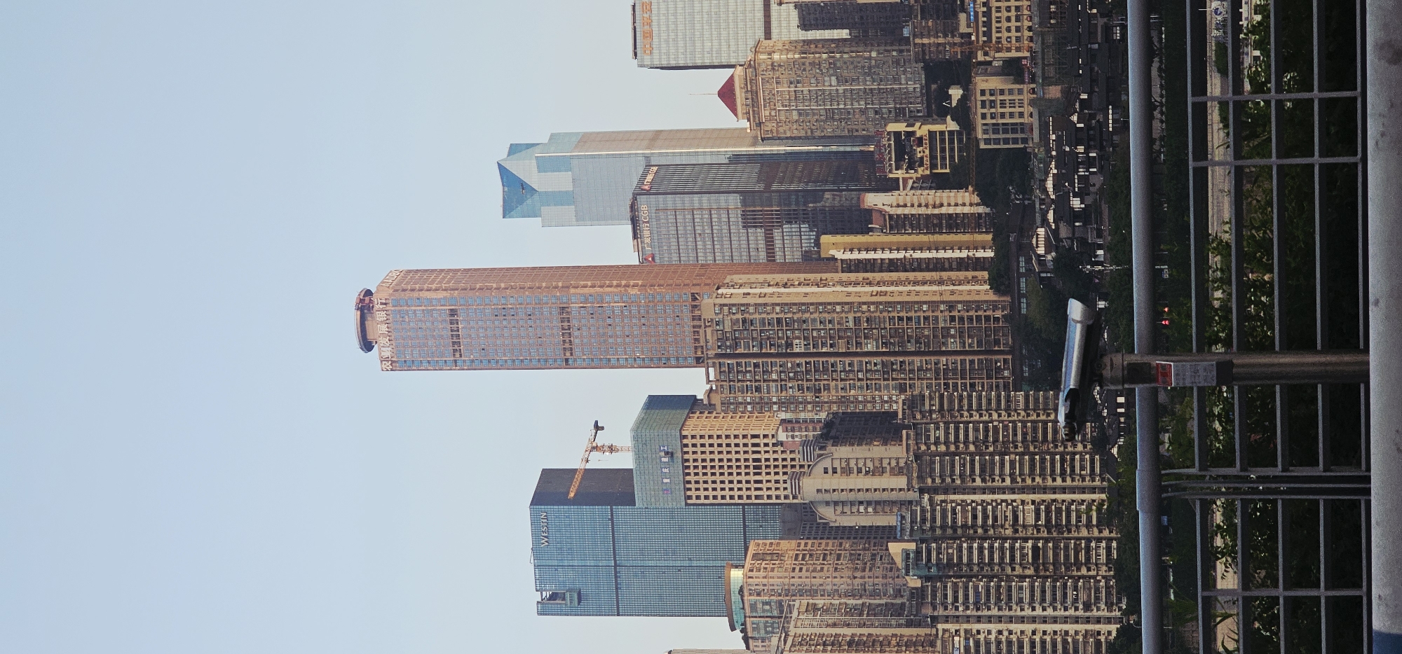This is a vertical, eye-level shot of the Chongqing skyline in Nan'an District, China, at dusk. The foreground features a metallic railing and a telescope on a stand. Behind these, a dense cluster of high-rise buildings dominates the mid-ground and background. The buildings vary in height, architectural style, and color, with a mix of modern glass structures and older, more ornate buildings. The tallest building, a reddish-brown skyscraper with Chinese characters and the letters "ADBOC" on top, is centrally located. To its left, a blue glass skyscraper with "WESTIN" visible on its facade stands tall. Further buildings are visible to the right and in the distance, some with illuminated windows. The sky is a clear, pale blue, suggesting the sun has recently set. The overall impression is of a bustling, modern city with a rich architectural history.