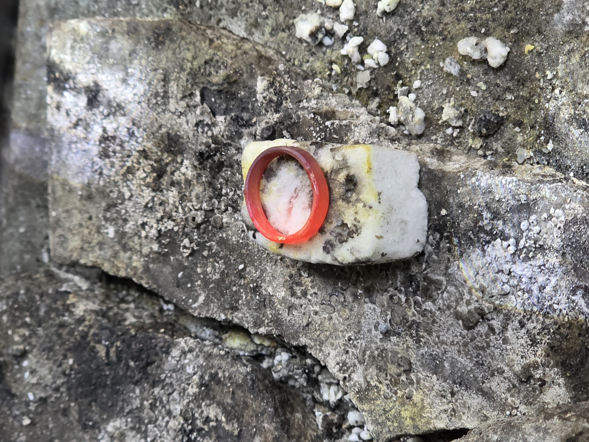 A close-up, overhead shot shows a single, bright red ring resting on a porous, white rectangular object. This object is placed on a rough, gray, textured surface that appears to be made of stone or concrete, possibly with some yellow and white mineral deposits. The background is a dark, uneven surface with visible cracks and small white particles. The lighting creates a slightly glossy sheen on the red ring. There is no indication of the city of Kazan, Russia, in the image.