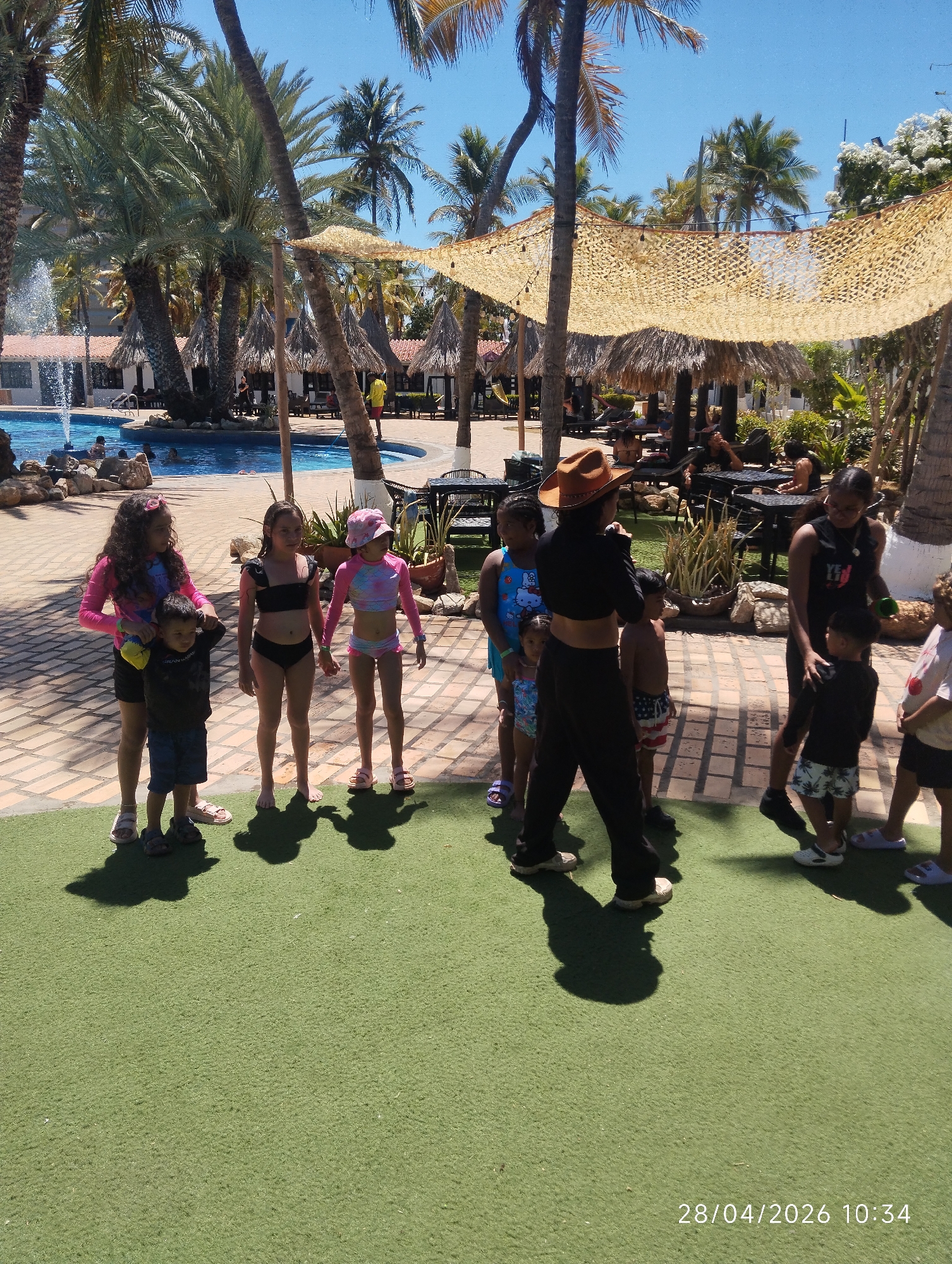 This media file depicts an outdoor scene at a tropical resort or hotel, likely in Porlamar, Venezuela, characterized by bright sunshine and a clear blue sky.
In the foreground, a group of approximately ten children and at least two adults are gathered on artificial green turf. One adult, wearing a brown hat and a black top and pants, stands centrally, seemingly leading or interacting with the children. Several children are dressed in various swimwear, including bikinis, long-sleeved swimsuits, and swim trunks, while others wear casual attire. Distinct shadows cast by the subjects on the turf indicate a late morning or midday sun.
The background showcases typical resort amenities. A large, irregularly shaped swimming pool with a fountain is visible, where several people, including children, are swimming or relaxing. Surrounding the pool area are paved surfaces and numerous tall palm trees, providing natural shade. Beneath a large, light-colored mesh shade structure and various thatched-roof cabanas, tables and chairs are set up, occupied by other guests relaxing or dining. Buildings with red-tiled roofs can be seen further in the background. The overall setting suggests a vacation or leisure environment.
The image conveys a relaxed, recreational atmosphere. The timestamp "28/04/2026 10:34" is visible in the bottom right corner, indicating the date and time the photograph was taken.