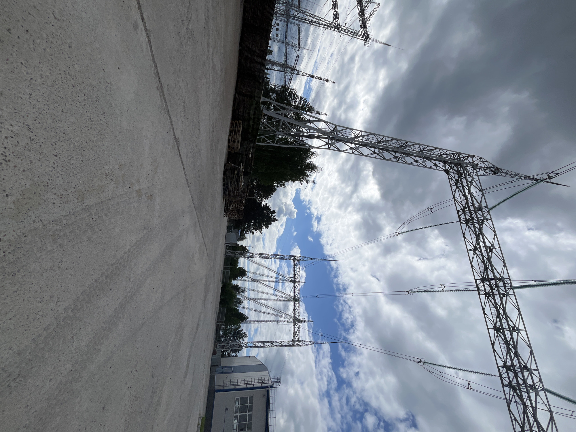 The image shows an outdoor scene featuring a concrete ground, a building, and multiple power line towers against a cloudy sky. The perspective is from ground level, looking upwards and across the scene.
In the foreground, there is a wide expanse of textured concrete. To the left and extending into the background, a low-rise building with grey and white panels and a row of windows is visible. Beyond the building and the concrete, there are numerous tall, metallic power line towers. These towers are constructed of intricate latticework and support thick electrical wires that stretch across the frame. Trees are visible behind the power line towers, suggesting a somewhat industrial or utility setting bordered by nature.
The sky is filled with dramatic clouds, with patches of blue sky visible between them. The lighting suggests it is daytime, possibly late afternoon or early evening, given the intensity of the clouds and the overall light. The weather appears to be overcast with potential for rain, indicated by the dark, heavy clouds.
There are no people or animals visible in the image. The overall mood is one of industrial infrastructure and the power of nature, with the imposing structures of the power lines contrasting with the dynamic sky. No visible text is present in the image. The location context provided is Chaikovsky, Russia, which aligns with the presence of such industrial infrastructure.