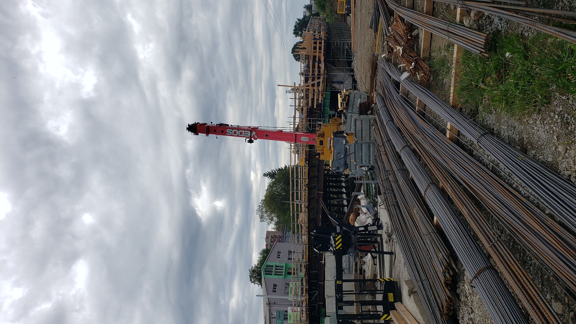 The image depicts a construction site in the city of Prešov, Slovakia, with a focus on bridge construction. A large red and yellow mobile crane, identified with the "VEDOS" logo, is prominently positioned. Numerous bundles of steel rebar are neatly arranged on the ground in the foreground and mid-ground. The construction site features wooden scaffolding and concrete structures that form the framework of what appears to be a bridge under development. In the background, buildings and trees suggest an urban or suburban setting. The sky is overcast with heavy clouds. No people are visible in the image, and the scene is static, indicating ongoing construction work.