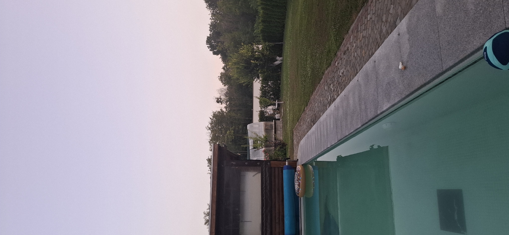 The image captures a serene backyard scene, likely in the city of Pleven, Bulgaria, at dusk. The focal point is a swimming pool with clear turquoise water, a portion of which is visible on the left and bottom of the frame. Floating in the pool is a donut-shaped inflatable ring. Adjacent to the pool is a paved walkway made of grey stone, bordering a lush green lawn dotted with white clover. Beyond the lawn, a dense line of trees forms the background, with a few structures, possibly greenhouses, visible amongst them. A wooden-roofed structure, likely a pool house or gazebo, is situated to the left. The sky is a soft gradient of pale purple and pink, indicative of twilight. No people are present in the scene.