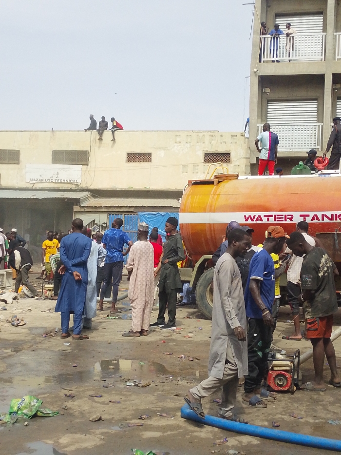 This image captures an active, unpaved urban outdoor scene in Fagge C, Nigeria, under a bright, clear sky.
A large, orange cylindrical water tanker truck, bearing the partially visible text "WATER TANK" on its side, occupies the mid-ground, positioned slightly to the right. To the rear and left of the tanker is a light-colored, single-story building with "MAZAF UTC TECHNICAL" signage and partially legible "COMPLE REVE" text. Three males are seated on the parapet of this building's rooftop. To the far right, a multi-story grey building features balconies with white railings and closed horizontal blinds; several males are visible standing on these balconies. At least two males are standing on top of the water tanker itself.
On the ground level, a crowd of approximately 30-40 individuals, predominantly males, are present. They wear a mix of traditional Nigerian attire (various colored kaftans, including blue, beige, and light grey) and casual Western clothing (t-shirts, trousers, shorts). A male in a light grey kaftan walks across the foreground from left to right. A small, red and black portable pump is visible on the ground near the front tire of the water tanker, surrounded by a group of males. A thick blue hose runs horizontally across the lower foreground. The ground surface is unpaved, appears wet with visible puddles, and is strewn with significant amounts of litter and debris, including plastic bags.