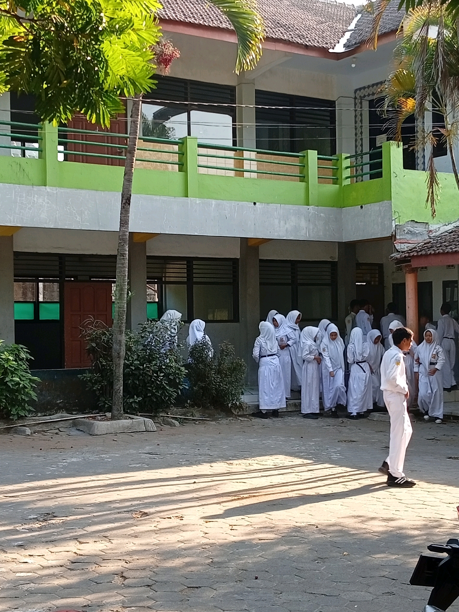 In Lawang, Indonesia, a group of students in white uniforms and headscarves are gathered in the courtyard of a school building. The building has a light green facade and brown doors. Sunlight casts shadows across the patterned ground. Some students are standing in small groups, while others are walking. One student in the foreground is facing away from the camera, looking towards the other students. The scene appears to be a normal school day.