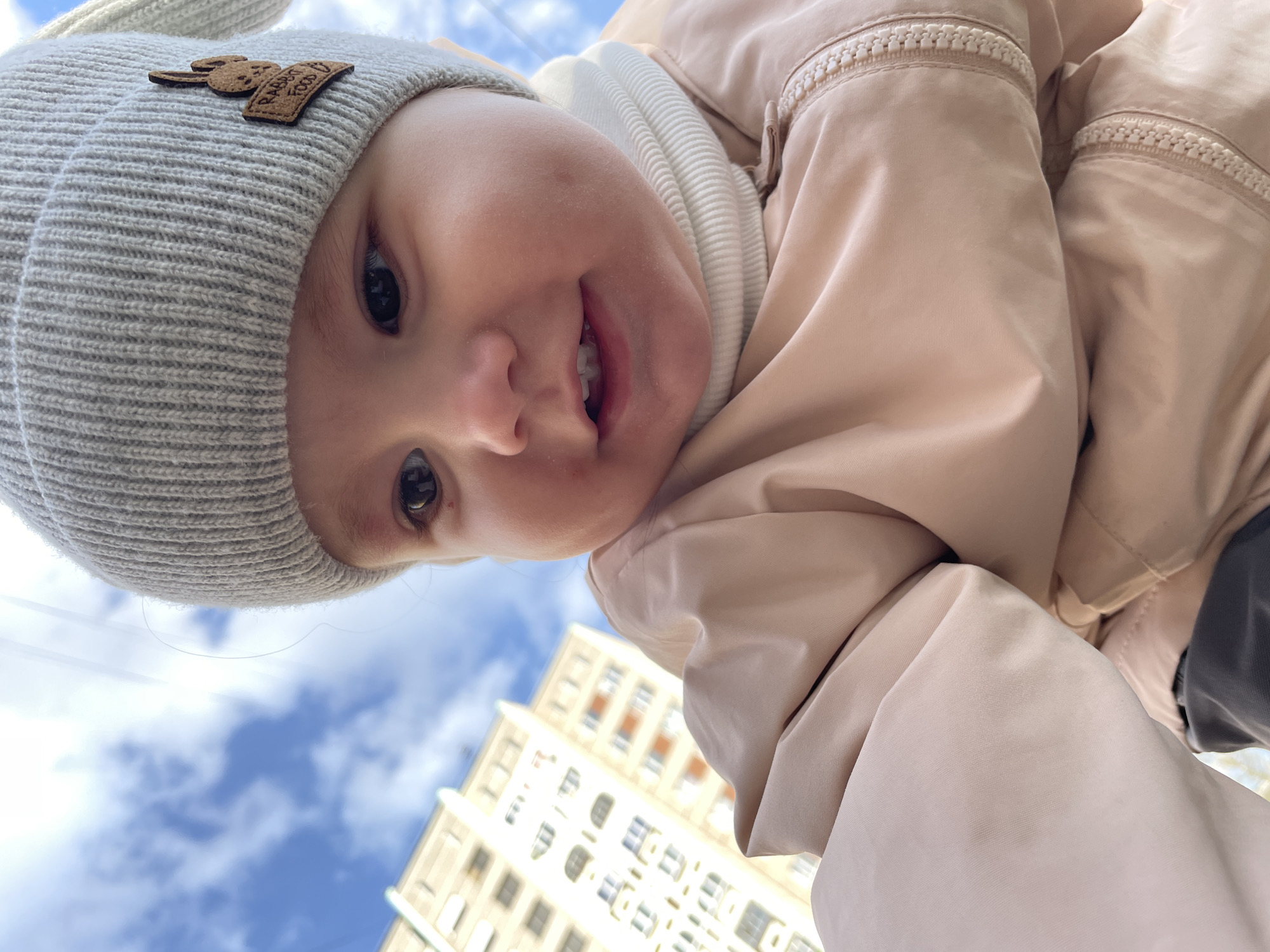 A close-up, low-angle shot shows a baby wearing a light gray knit hat with a brown patch depicting a rabbit and the words "Rabbit Food". The baby is also wearing a light pink puffer jacket and a white ribbed turtleneck sweater. The baby is smiling, revealing upper teeth. Behind the baby and slightly out of focus are a blue sky with scattered clouds and a multi-story building with numerous windows. The location is Izhevsk, Russia.