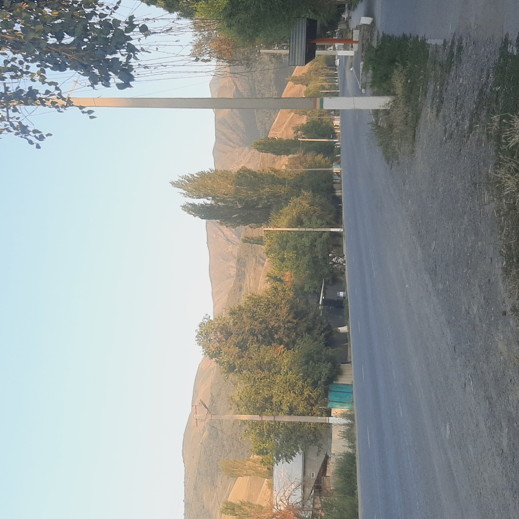 The image depicts a road with trees and mountains in the background, located in Sosnovka, Kyrgyzstan. The road is paved and has yellow lane markings. On the left side of the road, there are houses, fences, and trees. On the right side, there are more trees, a bus stop or shelter, and a sidewalk. The mountains in the background are rolling and appear to be dry. The sky is clear and light blue. There are a few poles with wires visible along the road. There are no people clearly visible in the image, though there might be some figures in the distance near the trees on the left. A white car is visible in the distance on the road. The overall scene suggests a rural or semi-rural setting.