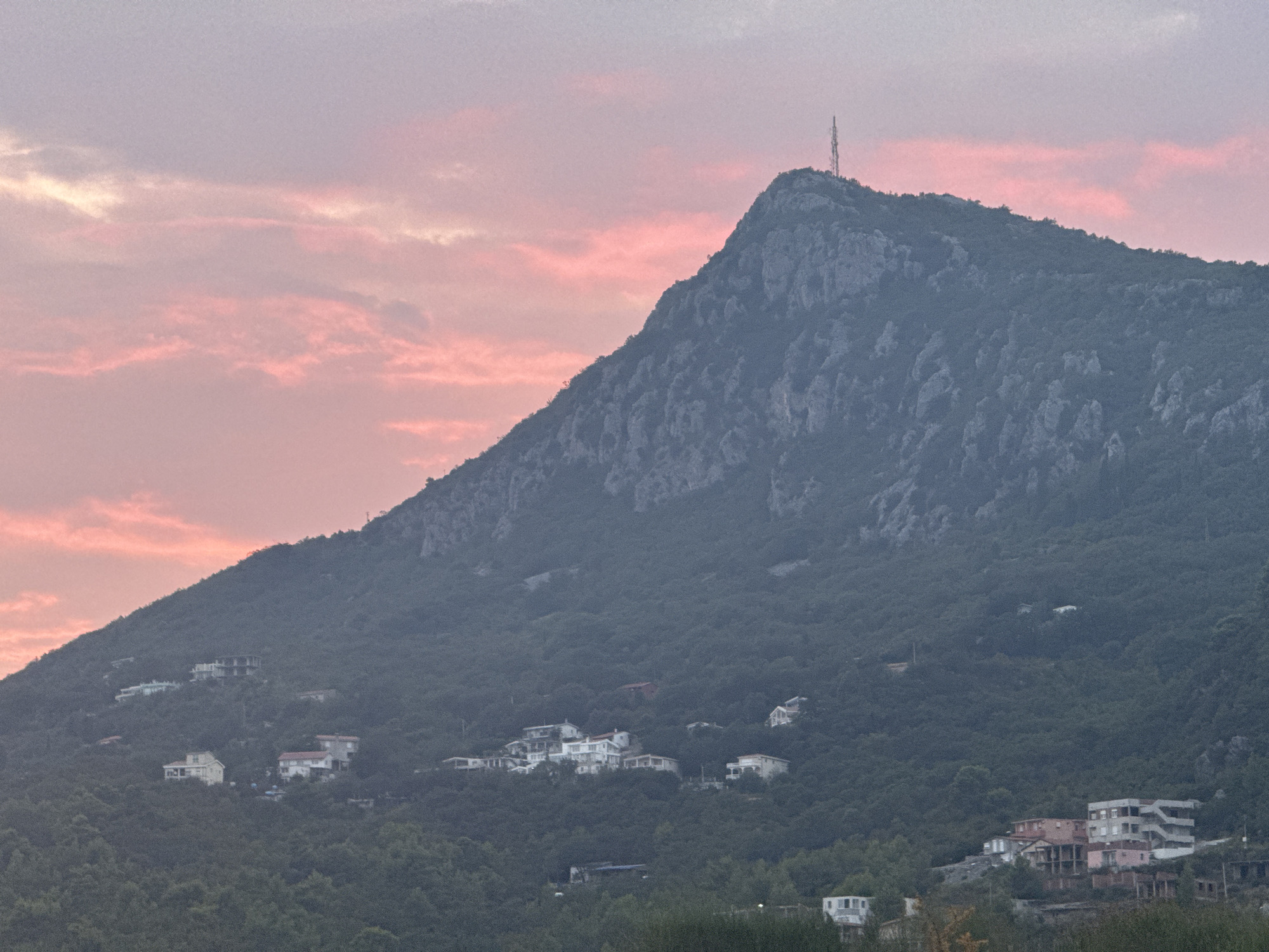 The image shows a mountainous landscape at dusk, with the sky painted in hues of pink and purple. The slopes of the mountain are covered in dense green foliage, with scattered buildings indicating a human settlement. A telecommunications tower stands prominently on the summit of the mountain. In the foreground, more buildings, some of which appear to be under construction or in disrepair, are visible nestled amongst the trees. The scene suggests a view from a vantage point overlooking the city of Sutomore, Montenegro, with its characteristic coastal mountain backdrop. The lighting suggests it is either sunrise or sunset.