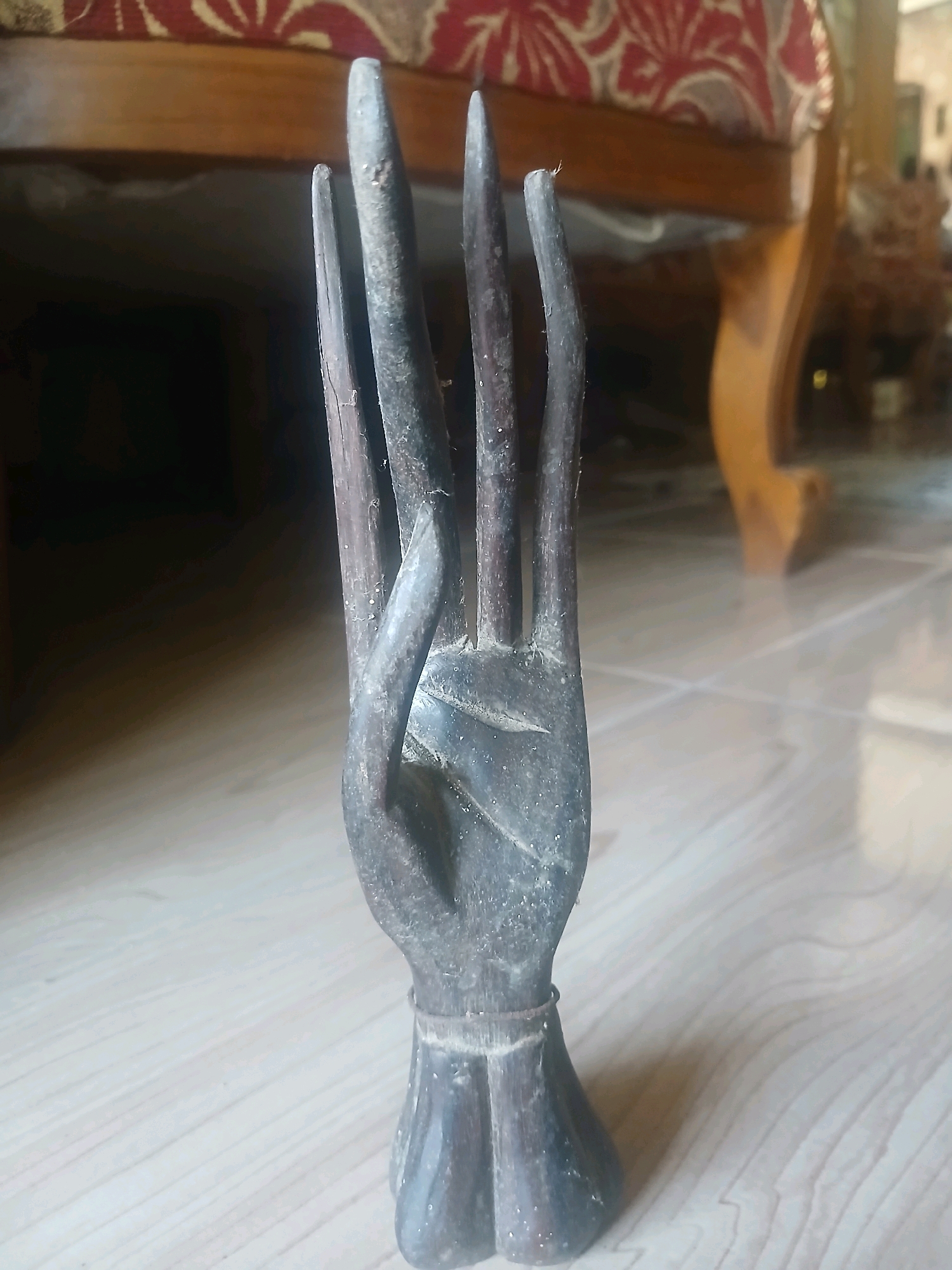 The image shows a close-up, eye-level, indoor shot of a dark-colored, wooden carved hand figurine standing upright on a tiled floor. The figurine is positioned in the foreground, with its four elongated fingers extended upwards and slightly curved. The hand itself appears to be in a gesture, possibly meditative or symbolic. The base of the figurine is flared, with what looks like a thin band of metal or dark material encircling it. The background is out of focus but reveals the legs of a wooden furniture piece with a patterned fabric seat, possibly a chair or sofa, and a plain tiled floor. The lighting suggests an indoor environment, likely a room in a house. This scene is likely located in Banyuwangi, Indonesia, as indicated by the user's prompt.