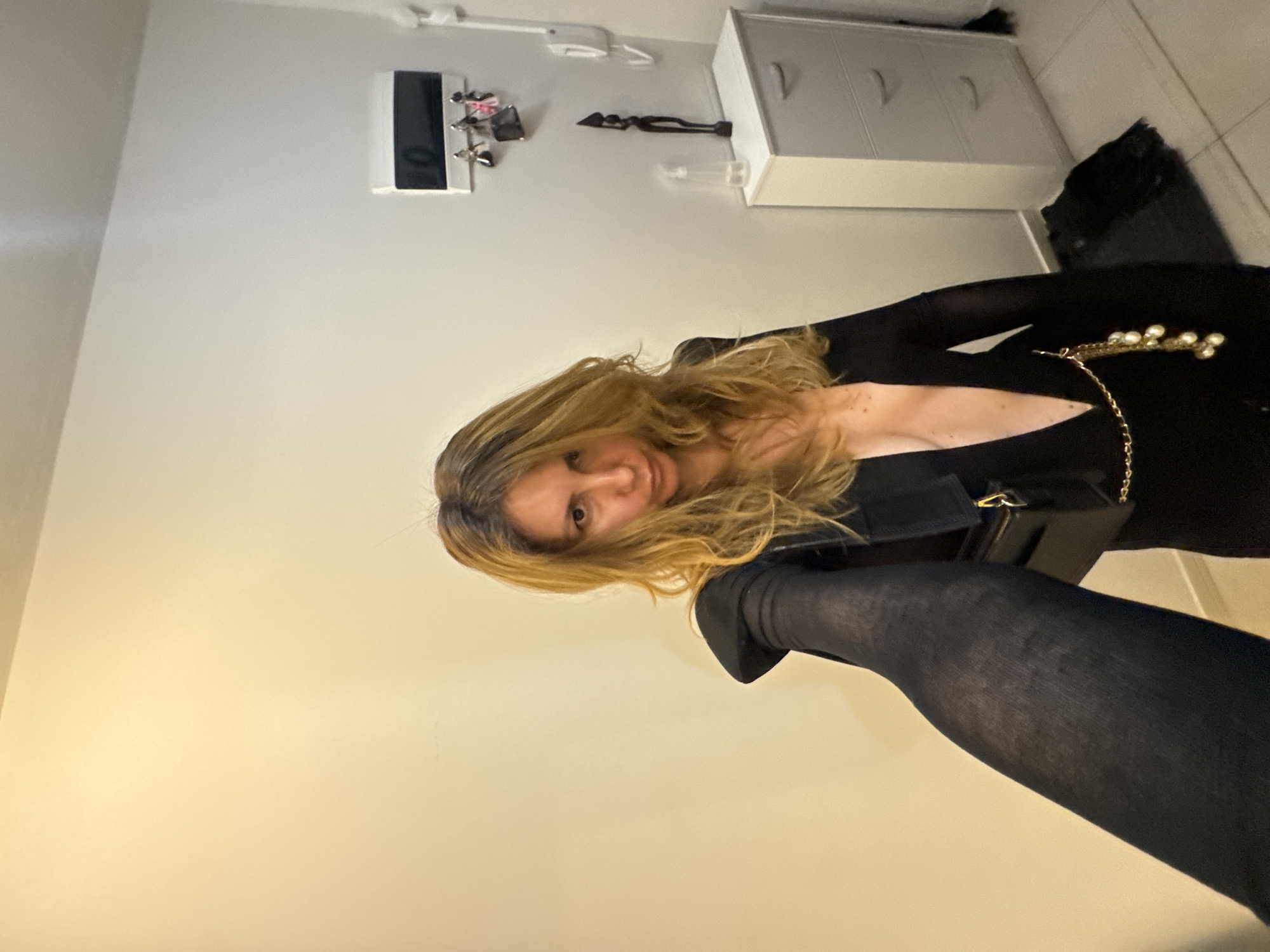 A woman is standing in what appears to be a hallway or entryway inside a building. She is wearing a black, long-sleeved, deep V-neck jumpsuit with a gold chain belt adorned with pearls. She has long, wavy blonde hair and is looking at the camera with a neutral expression. Her left arm and leg are prominently featured in the foreground, the arm is extended as if holding a camera for a selfie.
In the background, there is a light-colored wall on the left and a white wall on the right. Mounted on the white wall is a black and white rectangular item that appears to be a mail organizer or key holder, with several keys and small bags hanging from it. Below this, there is a slender dark wooden figurine standing on a white cabinet with grey drawers. To the right of the cabinet, a white wall-mounted telephone is visible. The floor is tiled in a light color. The lighting in the scene is dim and warm, likely from artificial sources. There is no visual information available to determine the location within the city of Sofia, Bulgaria.