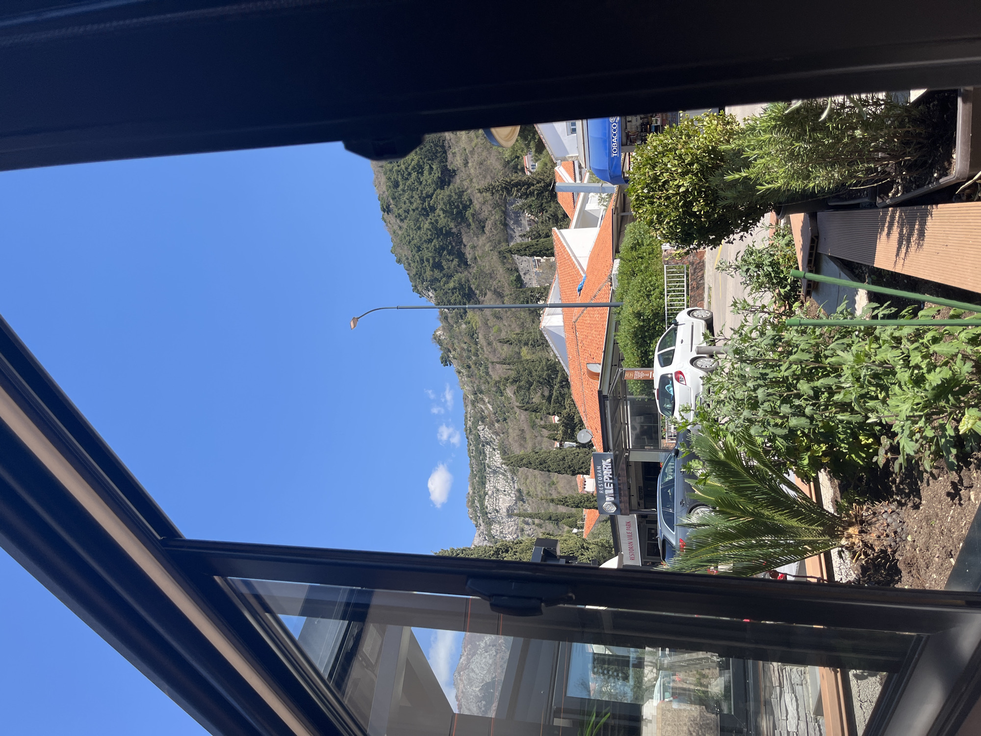 View from inside a building, through a window opening, across a street in Budva, Montenegro. Mountains rise in the background under a blue sky with scattered clouds. A row of buildings with red tile roofs are visible along the street. Street-level businesses are present, including a tobacco shop, and a restaurant/bar called "Vile Park" which is advertised on a sign. White passenger cars are parked on the street. Potted plants, bushes, and a small palm tree line the edge of the street.