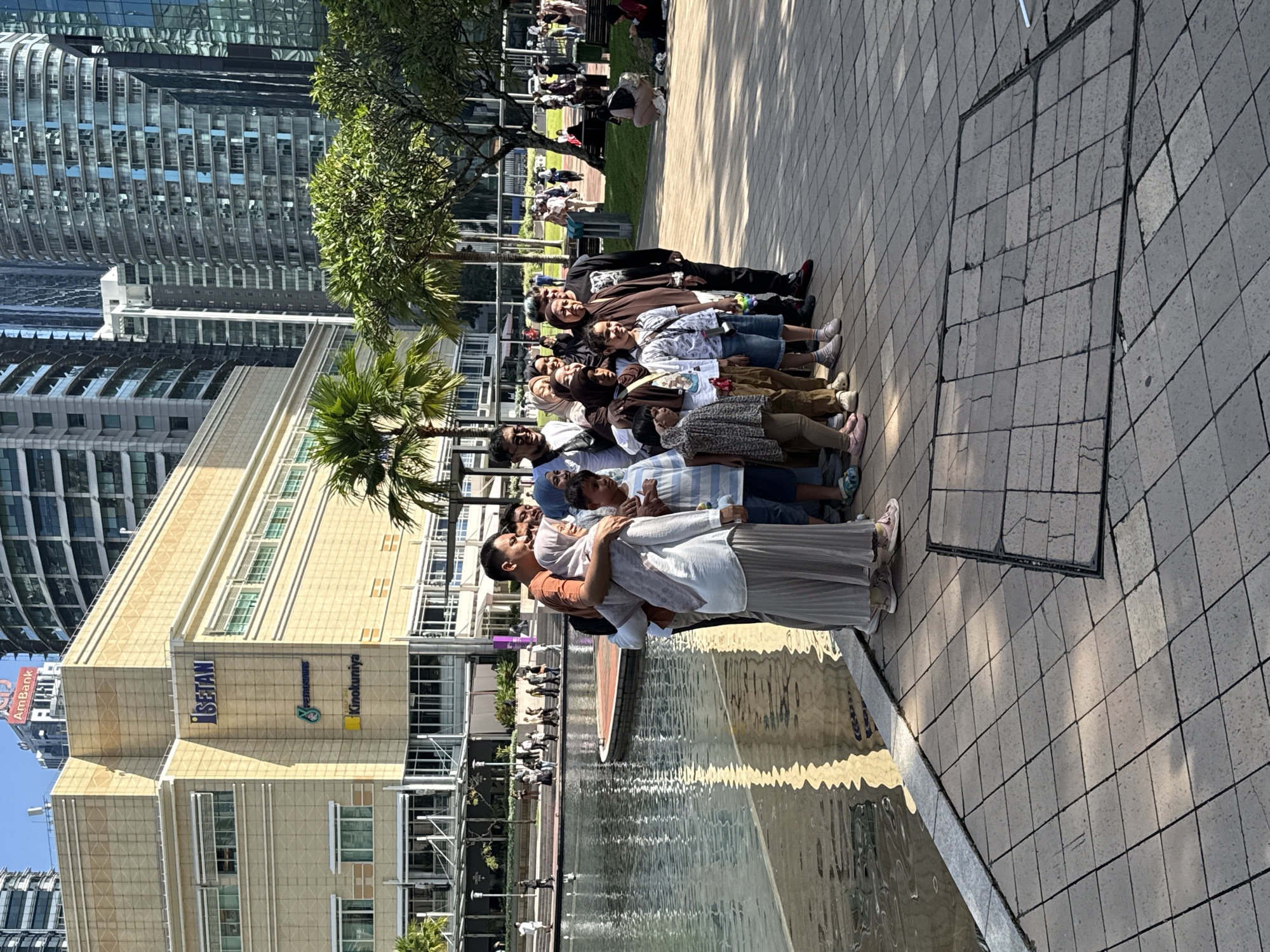 The photograph captures a daytime scene in Kuala Lumpur, Malaysia. A group of approximately 13 people of varying ages stands in a line on a paved area with tiled ground. They appear to be posing for a photograph. In the background, there is a water feature, likely a fountain or pool, with the reflection of buildings visible in the water. Behind the water feature, there is a multi-story building with signage for "ISETAN," "PARKSONS," and "Kinokuniya," indicating the presence of retail establishments. Lush green trees and palm trees are scattered around the area. Tall modern skyscrapers are visible in the distance, further suggesting an urban setting. The weather appears sunny and clear.