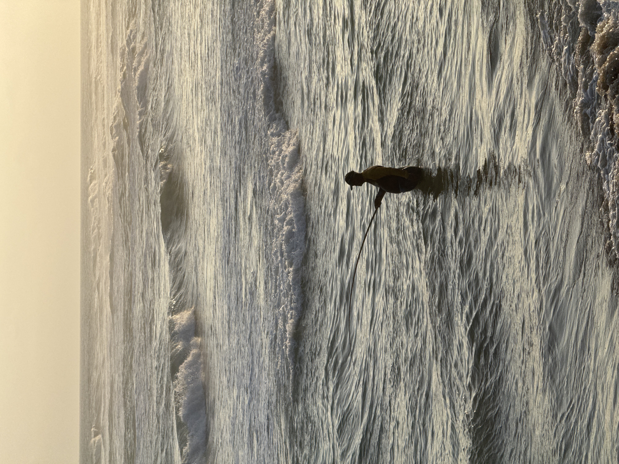The image captures a surfer silhouetted against the ocean's surface, likely during sunset, given the warm, soft light bathing the scene from the left. The surfer, a lone figure, is positioned in the water, holding a paddle and appearing to be either paddling or preparing to catch a wave. The waves themselves are the dominant element, creating a textured, dynamic surface with their crests and troughs. The water is a mix of white foam and deeper blues, reflecting the muted tones of the sky. The lighting suggests a tranquil atmosphere, with the sun low on the horizon casting a gentle glow and creating subtle shadows. The location is specified as Ahangama, Sri Lanka, a region known for its surf breaks, which aligns with the presence of a surfer in the water. There is no visible text in the image.