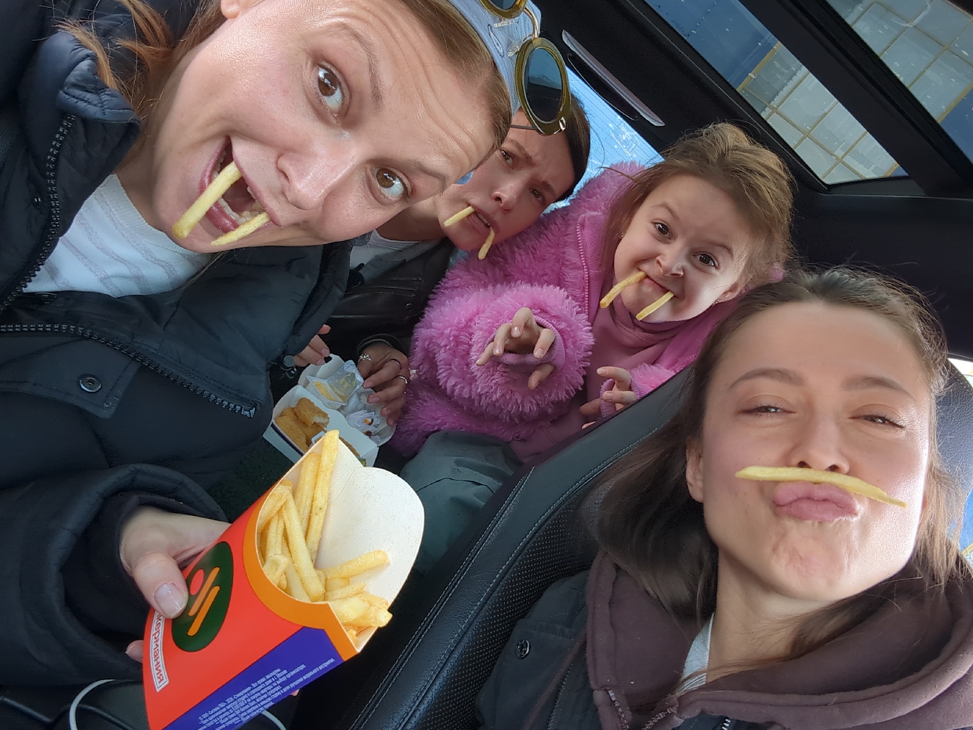The scene depicts the interior of a vehicle in Golikovo, Russia. Four females of varying ages are present, positioned in the vehicle's cabin. In the immediate foreground, a woman with light brown hair, wearing a dark puffer jacket over a light-colored striped top, looks towards the camera with her mouth open, from which two french fries protrude. Her right hand holds a red and orange fast-food carton containing french fries, displaying the brand "ВКУСНО и ТОЧКА." Behind her, partially visible, another woman wearing a dark jacket has two french fries positioned in her mouth; a pair of sunglasses rests on her head. Adjacent to her, a young girl in a pink fluffy jacket also has two french fries extending from her mouth. In the background, a fourth woman, wearing a brown hooded sweatshirt, poses with a single french fry horizontally placed above her upper lip, with her lips puckered. An additional fast-food container, possibly holding chicken nuggets, is visible to the left. A vehicle sunroof is visible in the upper left portion of the frame.