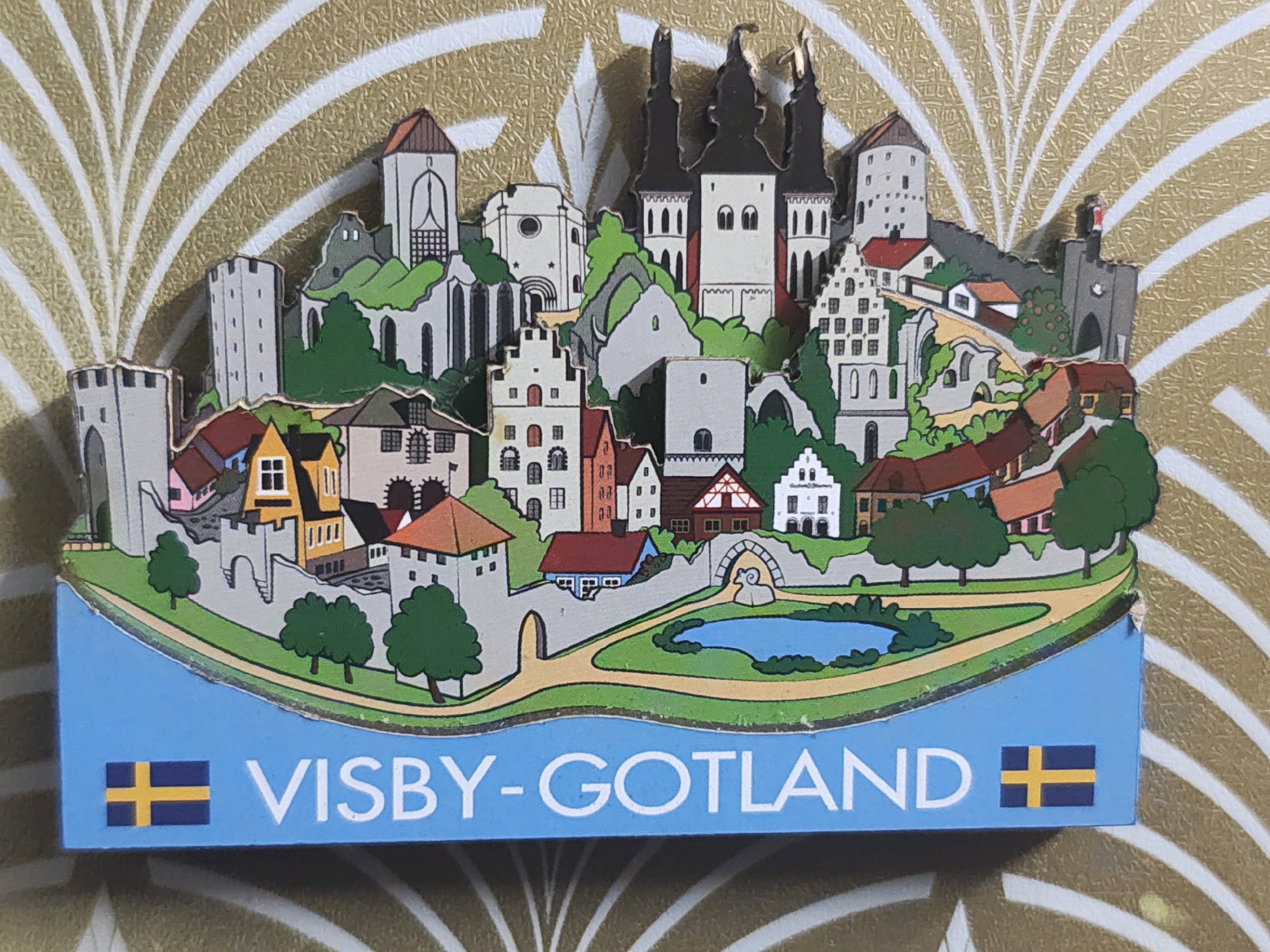 The image shows a souvenir magnet depicting a cityscape. The magnet features a cluster of stylized buildings with varying architectural styles, including towers, spires, and rooftops. The buildings are arranged on a tiered landscape, with pathways and a small blue body of water. The magnet has a blue border at the bottom with the text "VISBY-GOTLAND" and two Swedish flags. The background of the magnet is a textured gold and white pattern. There are no people or explicit actions depicted in the image. The image does not show or relate to the city of Uzhavoor, India.
