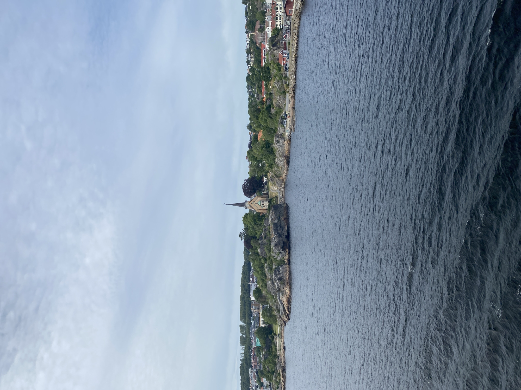 This is a landscape photograph taken from the water, looking towards a rocky coastline. The image is oriented vertically, with the sky at the left and the sea at the right. The coastline is dotted with buildings, many of which appear to be houses with red roofs, nestled among lush green trees. A prominent stone church with a spire stands on a cliff overlooking the water. The sea is a deep blue with visible ripples, suggesting a gentle breeze. The sky is overcast with soft, diffused light, indicating it is likely daytime, possibly late afternoon or early morning. The scene evokes a sense of tranquility and the picturesque beauty of a coastal town. The location is identified as Larvik, Norway, which aligns with the typical architecture and coastal landscape of Scandinavian regions. There is no visible text in the image.
