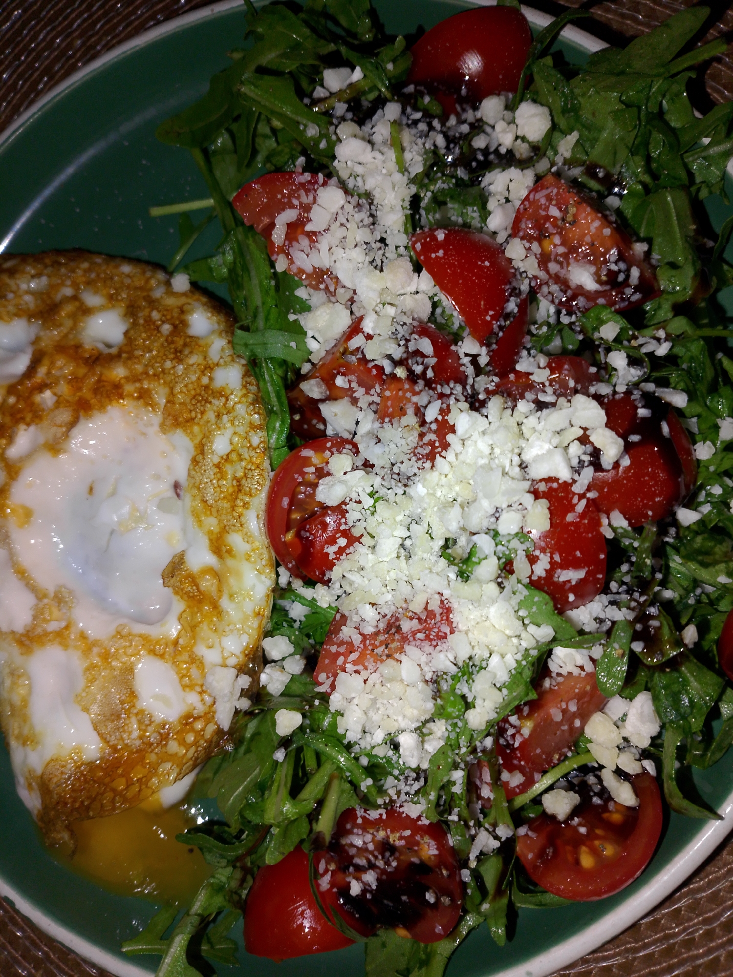 A top-down, close-up shot depicts a green plate containing a meal consisting of an over-easy fried egg and a salad. The egg has a cooked, golden-brown edge and a partially runny yolk at the bottom. The salad is a mix of arugula, halved cherry tomatoes, and crumbly white cheese. A dark balsamic glaze is drizzled over the salad. The background is a woven, possibly straw-like surface. There is no evidence that this image was taken in Izhevsk, Russia.