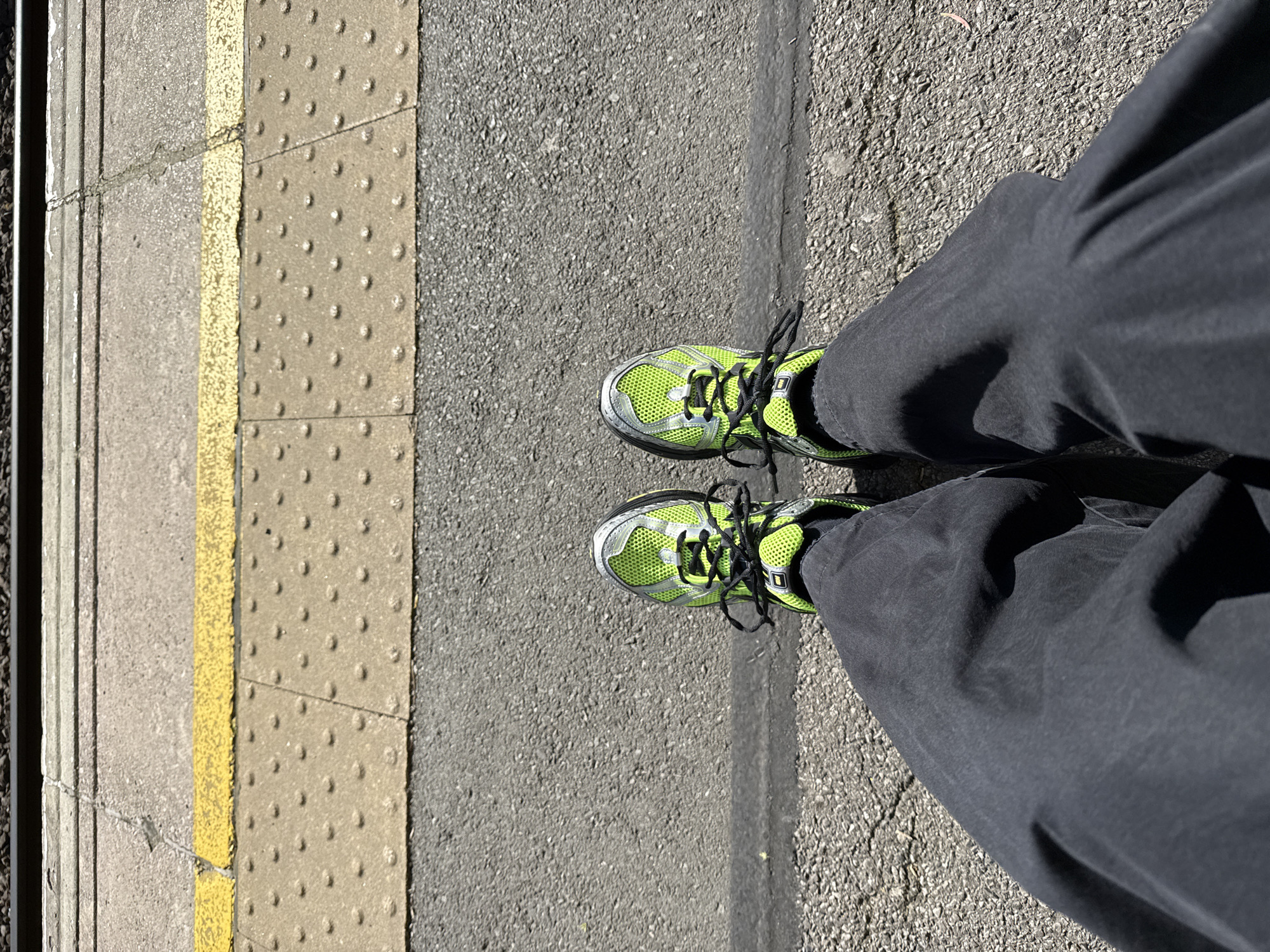 The image shows a pair of legs and feet standing on a train platform in London. The person is wearing grey trousers and bright green running shoes with black laces. The platform edge is visible, with a tactile paving surface indicating the proximity of the tracks. A yellow safety line is painted on the platform surface. The surface is grey concrete.