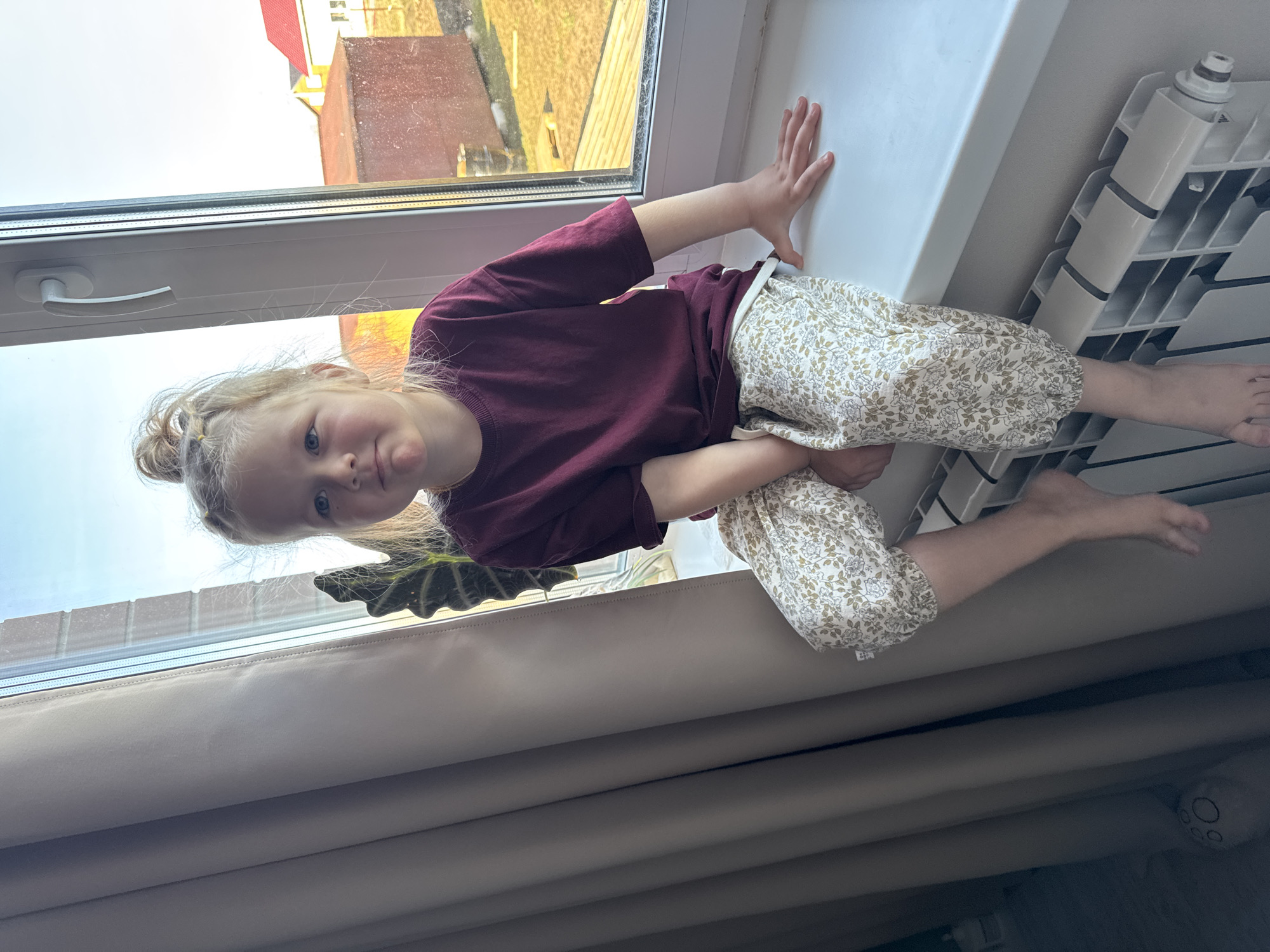 A young girl with blonde hair, tied in a bun, is captured in a sideways-oriented photograph, appearing to lie on a window sill. She gazes directly at the camera with a neutral, slightly inquisitive expression.
The girl is dressed in a plain maroon long-sleeved top and light-colored, loose-fitting pants featuring a subtle floral or leafy pattern. Her feet are bare, with one resting near a white radiator. Her hands are placed on the white window frame and the wall/sill next to the radiator.
Behind her head, a dark green plant leaf is partially visible. The window itself has a white frame and handle, looking out onto an outdoor scene. The exterior appears to be under soft, diffused daylight, possibly indicating an overcast sky or an early/late hour. Through the window, parts of a building with a reddish roof and a textured facade are visible. Below the window sill, cream-colored pleated blinds or curtains are drawn. The scene depicts an indoor setting, likely a home, with the child comfortably positioned by the window. There is no visible text in the image.