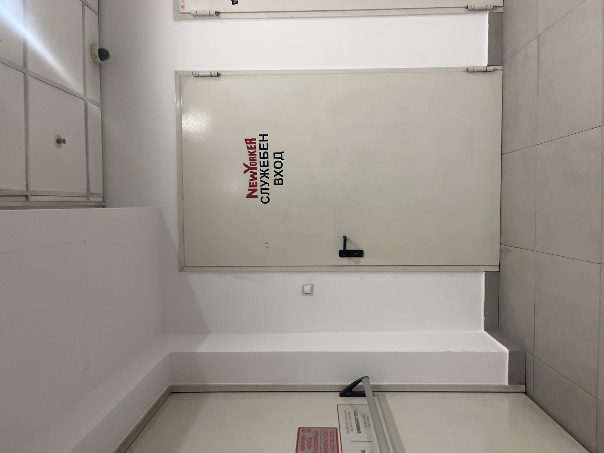 The image shows a white door with black and red text in Cyrillic. The text includes "NEW YORKER", "СЛУЖЕБЕН ВХОД". There is a black door handle and a small white switch on the wall below the door. The environment appears to be an interior space, likely a commercial building. Based on the Cyrillic text, it is likely located in a country that uses the Cyrillic alphabet, such as Bulgaria. Although there is no way to confirm if the photo was taken in Sofia, Bulgaria, it is probable given the description on the doors.