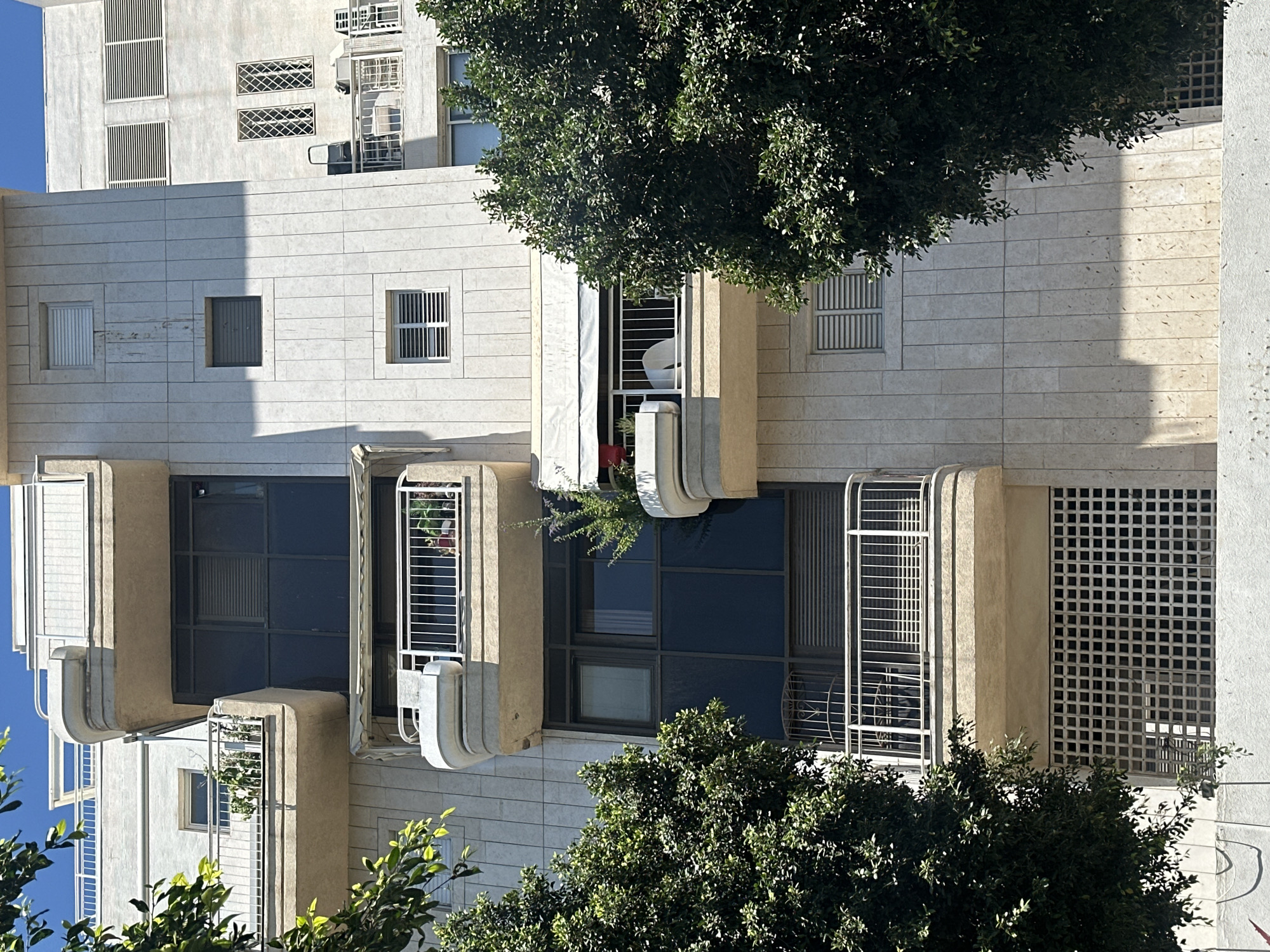 The image captures a portion of a modern apartment building in Tel-Aviv, Israel, bathed in sunlight. The building's facade is clad in light-colored stone tiles, arranged vertically. Various balconies punctuate the facade, some featuring white railings and planters with greenery. One balcony on the middle floor has a white awning extended.
On the right side of the building, a section with horizontal louvered windows is visible, along with two decorative latticed windows on an upper floor. Another section of the building, to the left and slightly set back, shows a balcony with plants and a white railing.
The lower part of the building features a prominent patterned grate. Lush green trees flank the building on both the left and right sides, their branches and leaves partially obscuring the lower levels. The sky above is a clear blue. No people are visible in the image. The overall impression is of a well-maintained urban residential structure in a sunny climate.