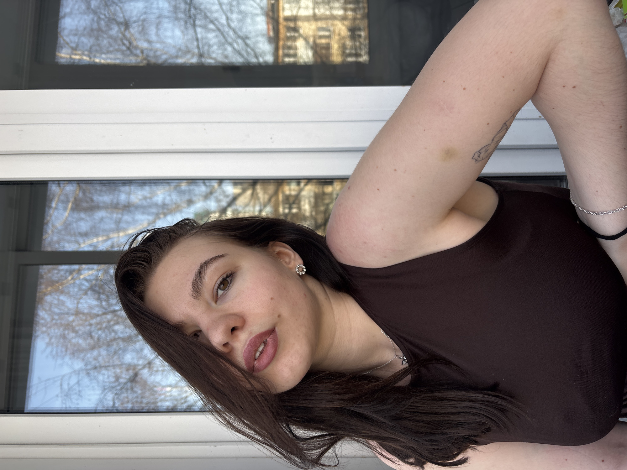 A woman is photographed indoors with light skin, dark brown hair, brown eyes, and wearing a brown top. The background includes a window with a view of buildings and trees, likely in an urban environment. The location is possibly Perm, Russia. The woman has a tattoo on her left arm, visible next to her shoulder, and a silver bracelet on her left wrist. Her hair partially covers her face.