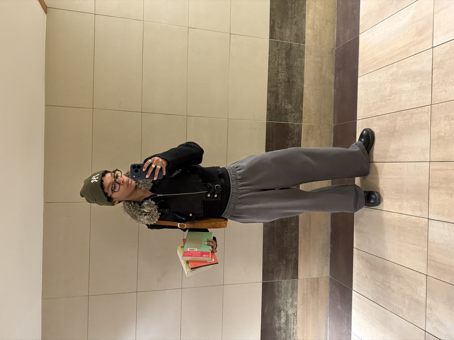 A person is depicted standing indoors, taking a mirror selfie in Champs-sur-Marne, France. The individual, wearing an olive green beanie with a white "NY" logo, glasses, a black cropped jacket with a fur-lined collar, a black undershirt, grey wide-leg sweatpants, and black shoes, holds a dark blue smartphone in their right hand, angled to capture their reflection. Two rings are visible on their right hand. In their left arm, they carry a stack of four books with variously colored spines (red, green, orange, light green/yellow). A brown shoulder bag hangs from their left shoulder. The background consists of a tiled wall featuring a main section of light beige/cream rectangular tiles and a lower horizontal band of darker, mottled brown/grey tiles. The floor is covered in light beige/cream square tiles arranged in a grid pattern.