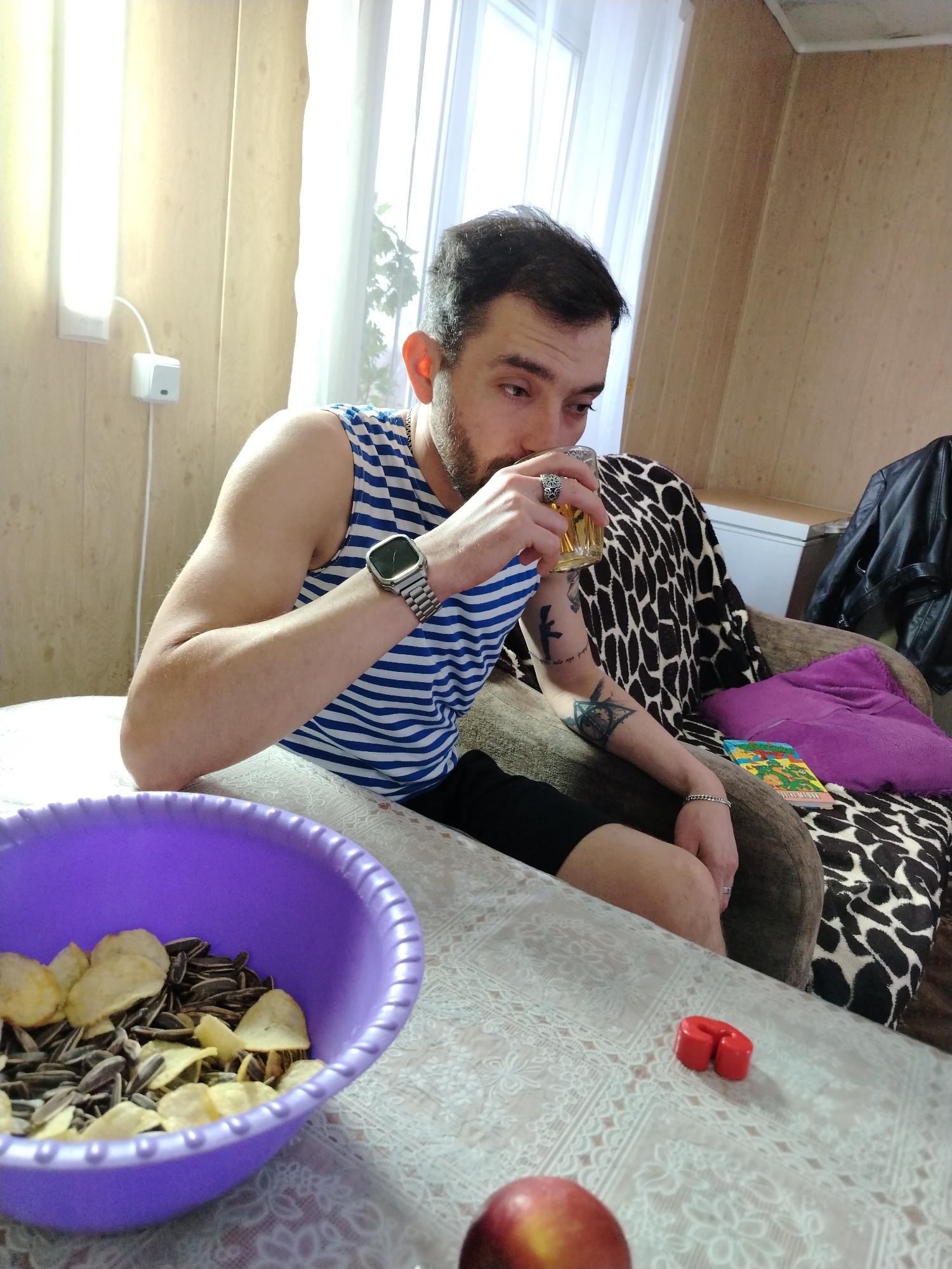 A man sits at a table in what appears to be a home interior. He wears a blue and white striped tank top, a wristwatch, and has tattoos on his forearm. He is drinking from a glass. On the table, there's a purple bowl filled with sunflower seeds and potato chips, a red object of unknown use, and an apple. A blanket with a black and white animal print is visible on the chair behind him. The location is identified as Большая Уса, Russia.