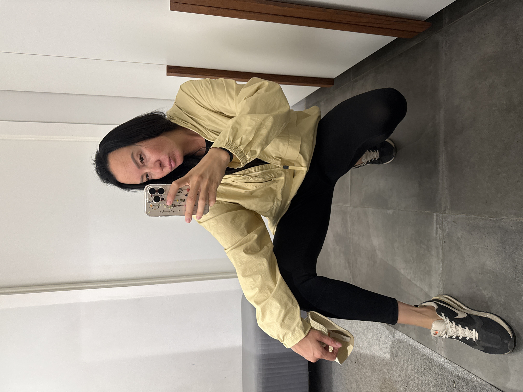 A woman is squatting in what appears to be a fitting room, holding a smartphone in her right hand and taking a selfie. She is wearing a light yellow hooded jacket zipped halfway, a black t-shirt underneath, black leggings, and dark colored athletic shoes with white laces. She is holding a folded piece of light-colored fabric in her left hand. The room has a tiled floor and white walls, with a gray upholstered bench on the left and wooden framed panels on the right. The overall impression is of a casual, indoor setting, possibly a retail store or changing area. There is no indication that this scene is located in Chaikovsky, Russia.