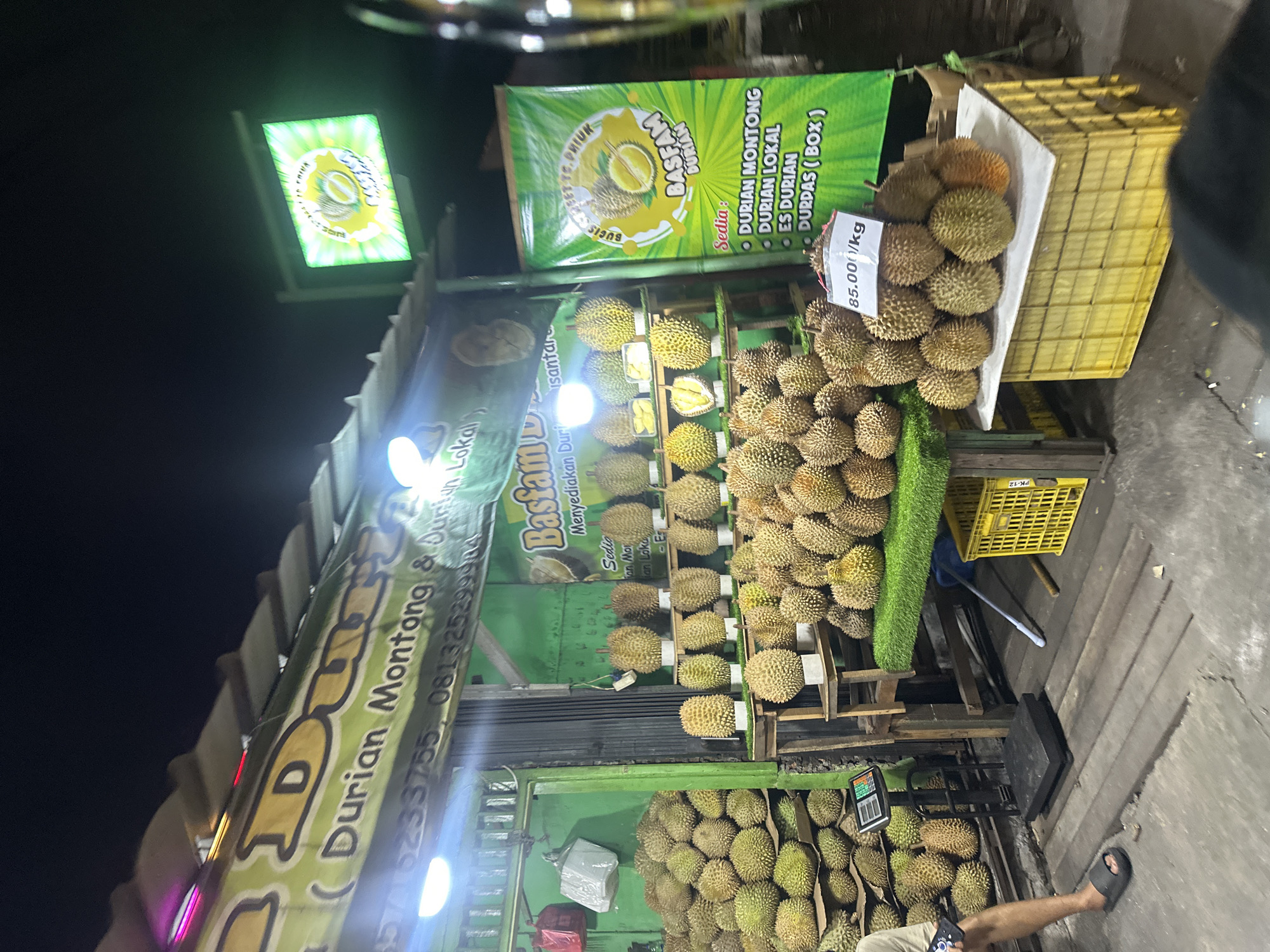 The image shows a durian fruit stand at night in North Jakarta, Indonesia. The stand is stocked with many durians, stacked in piles and on shelves. Signs advertise "Durian Montong" and "Durian Lokal" (local durian). One sign specifies the price as 85.000/kg. A partial view of a person's legs and feet is visible near the bottom of the frame.