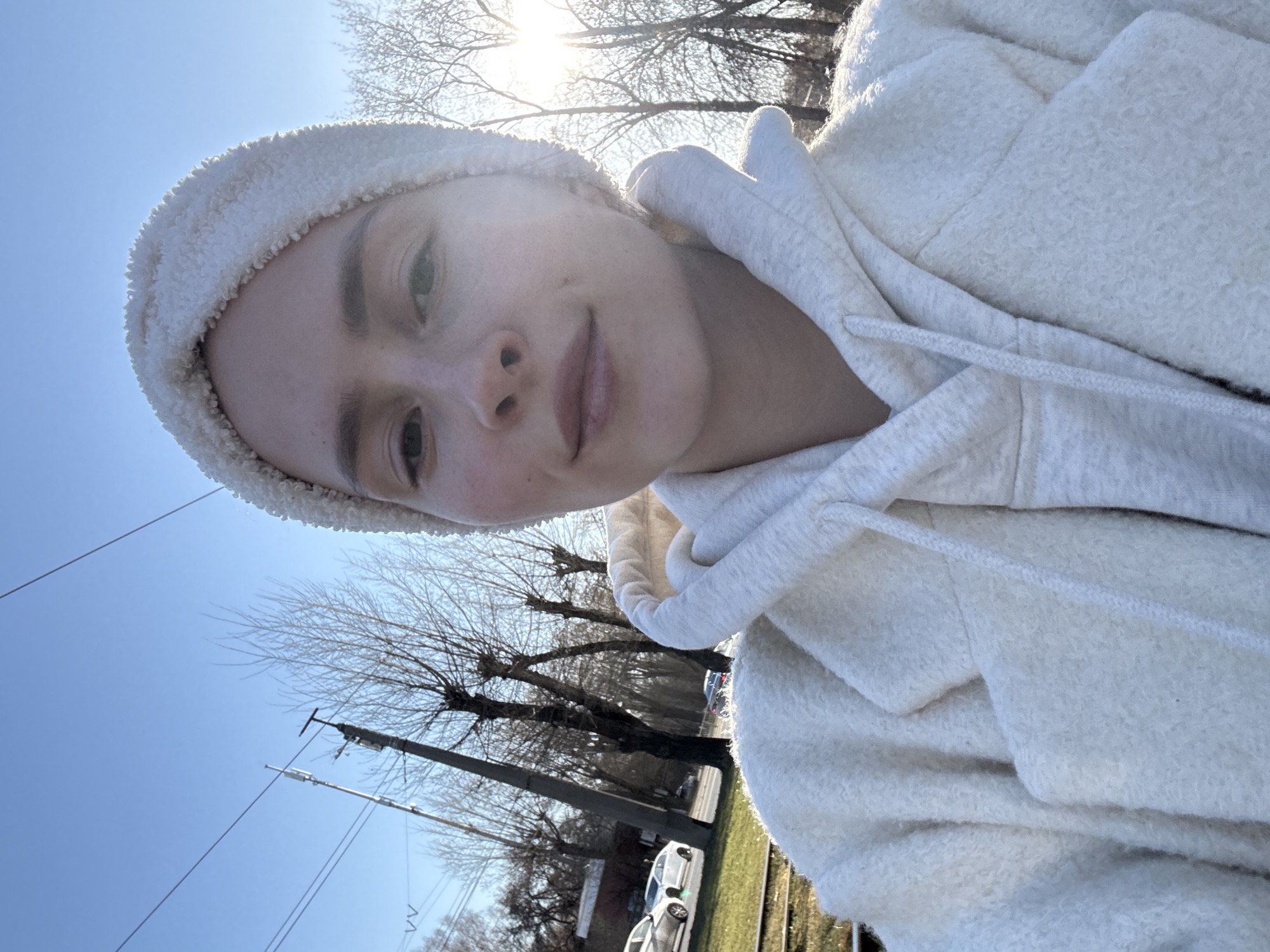 A woman wearing a white beanie and a white hooded jacket is taking a selfie outdoors. The location appears to be near a street with parked cars, power lines, and bare trees. The sky is clear and blue. The sun is shining brightly, possibly causing the overexposure in parts of the image. Without further information, a specific location in Yekaterinburg cannot be determined.