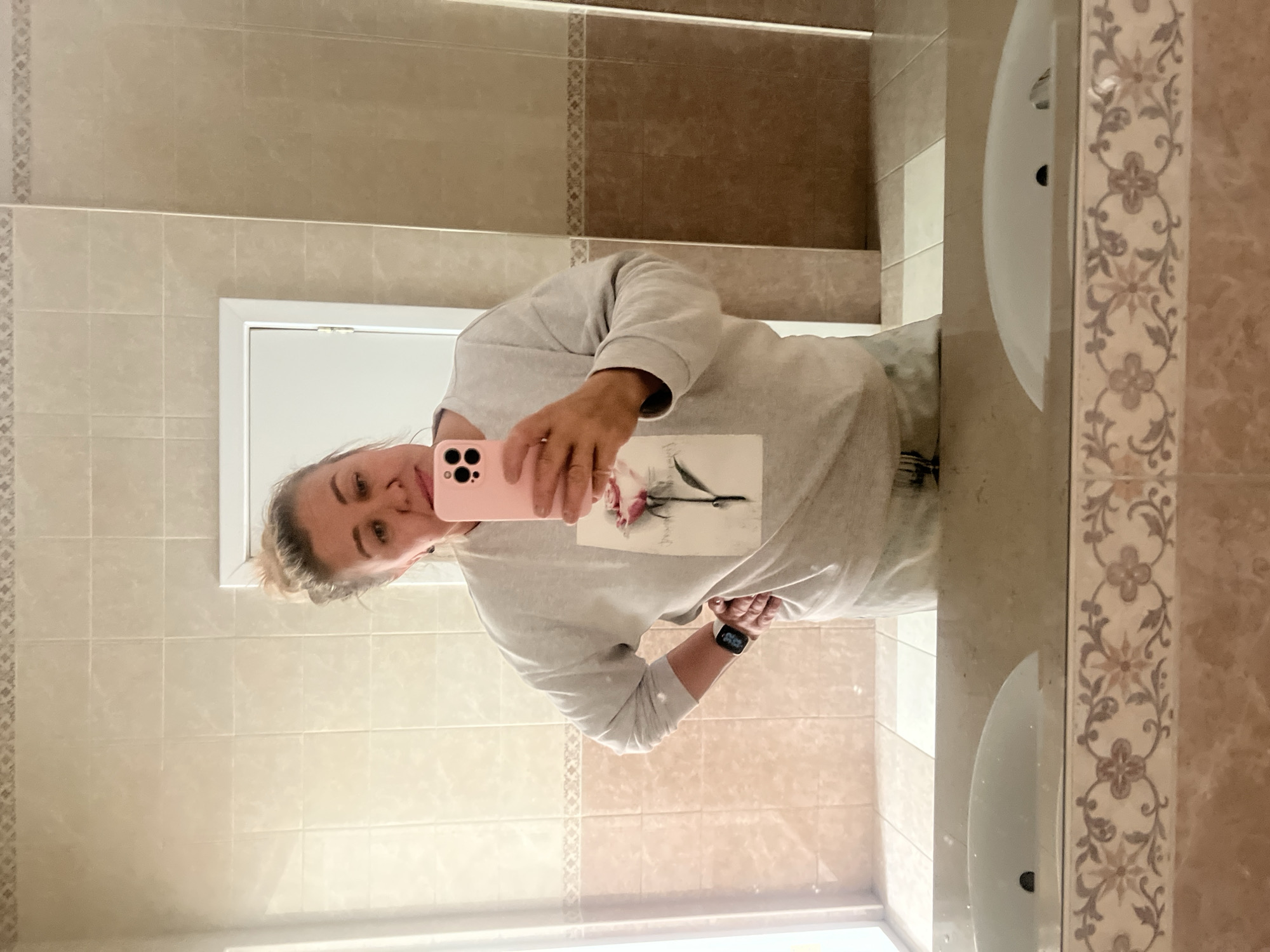 The image, captured as a mirror selfie and rotated sideways, features a woman posing in a bathroom. She has light-colored hair, likely tied back, and a neutral to slightly smiling expression. The woman is wearing a light gray long-sleeved sweatshirt that has a subtle graphic design of a flower and foliage on its front. On her left wrist, she wears a black smartwatch displaying "09:02". She holds a pink smartphone, with its triple camera array visible, in her right hand, capturing her reflection.
The bathroom's interior is visible in the reflection. The walls are tiled with square tiles in two main tones: lighter beige/cream on the upper and lower sections, and a darker brown/tan band in the middle, separated by a thin, decorative border tile. Two white ceramic sinks are partially visible in the reflection, integrated into a vanity that also features decorative patterned tiles with floral and geometric motifs. A white doorframe is seen in the background reflection. The scene depicts an indoor setting, and the provided context places this location in Votkinsk, Russia.