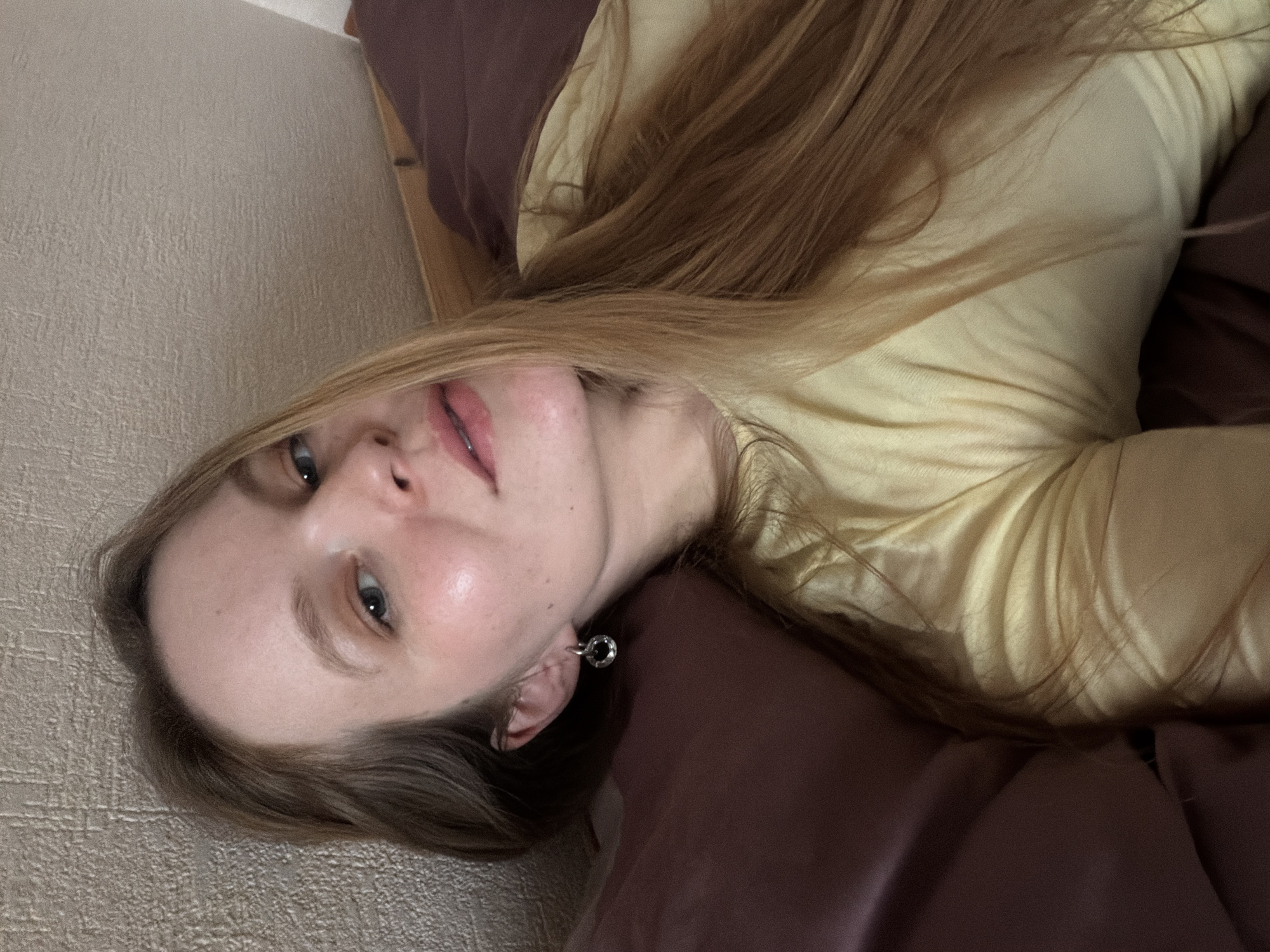 A young woman with long blonde hair is lying down. She is wearing a yellow top and appears to be on a brown pillow or cushion. Her head is close to a beige wall. The scene does not provide information to identify the location in the city of Izhevsk, Russia.