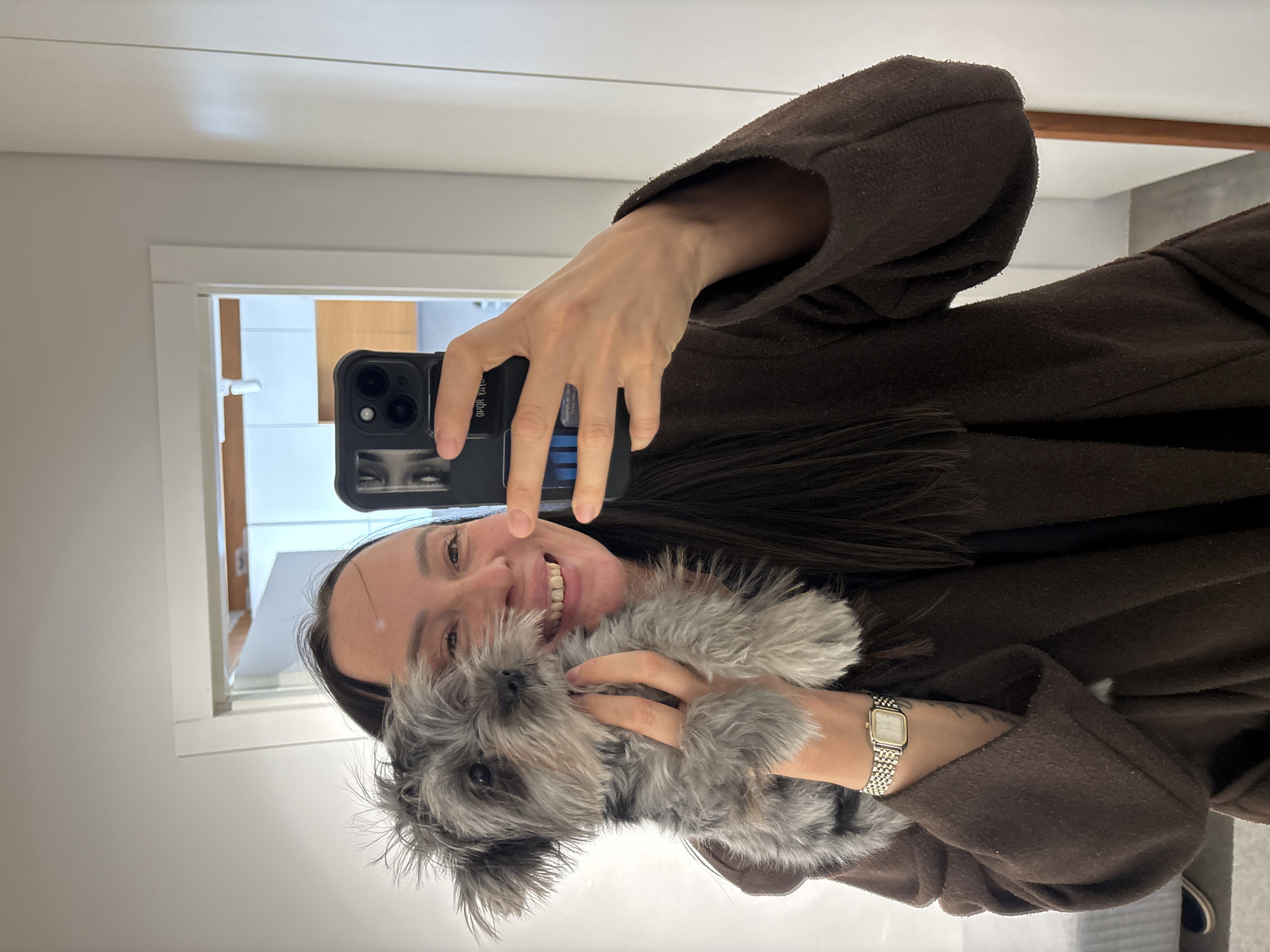 A woman is holding a gray Schnauzer-type dog while taking a selfie in a mirror. The woman is wearing a brown coat with a fur collar. She has dark hair and is smiling. She is holding a black iPhone with a design on the back. The background appears to be a bathroom with a white door and white walls. There is no information in the image to support the location being Chaikovsky, Russia.