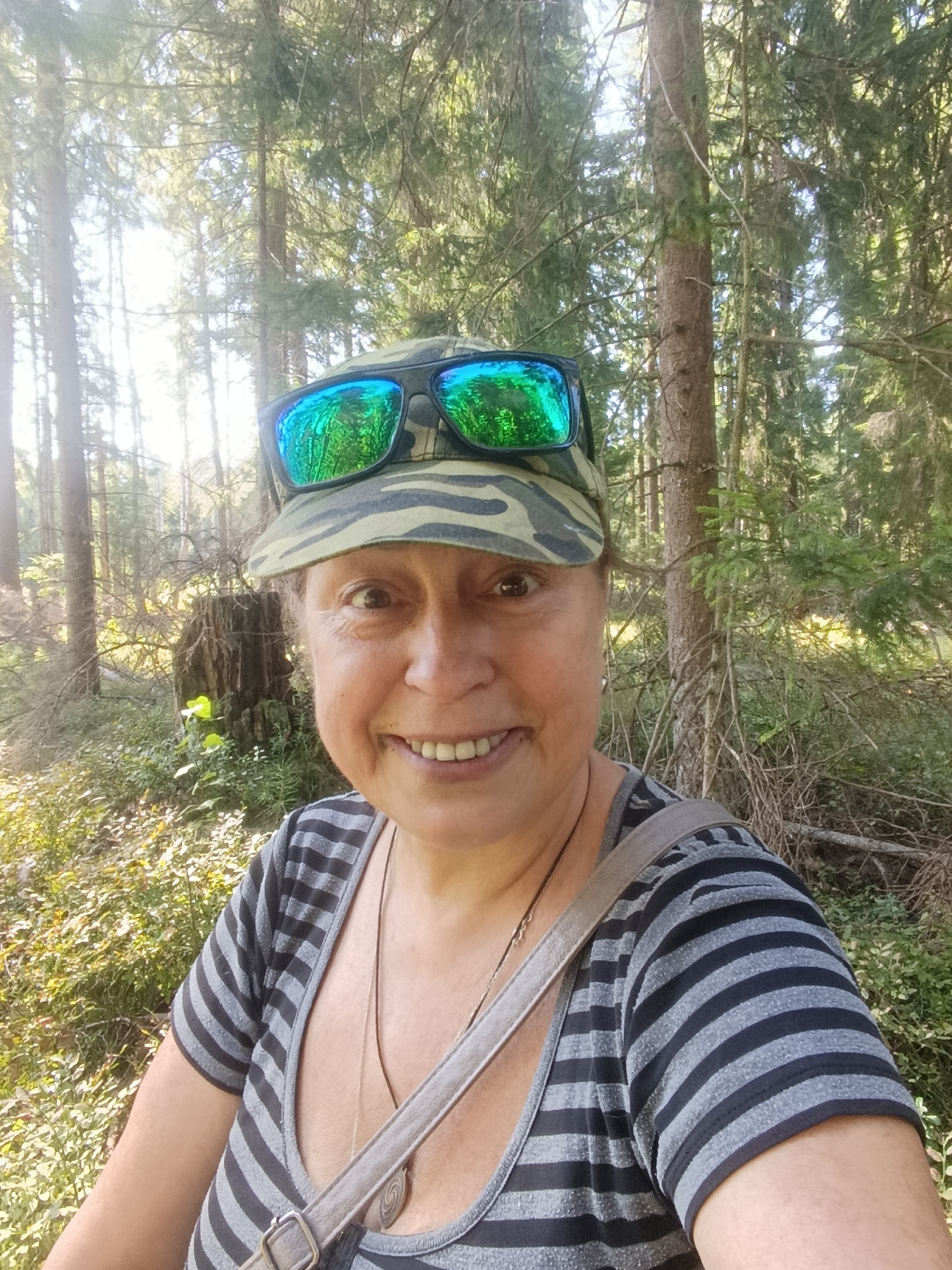 A woman is taking a selfie in a forest. She is wearing a camouflage baseball cap with sunglasses on top and a gray and black striped t-shirt. A bag strap is visible across her shoulder. The background shows a dense forest with tall trees, undergrowth, and sunlight filtering through the leaves. The location is identified as Gribchikha, Russia.