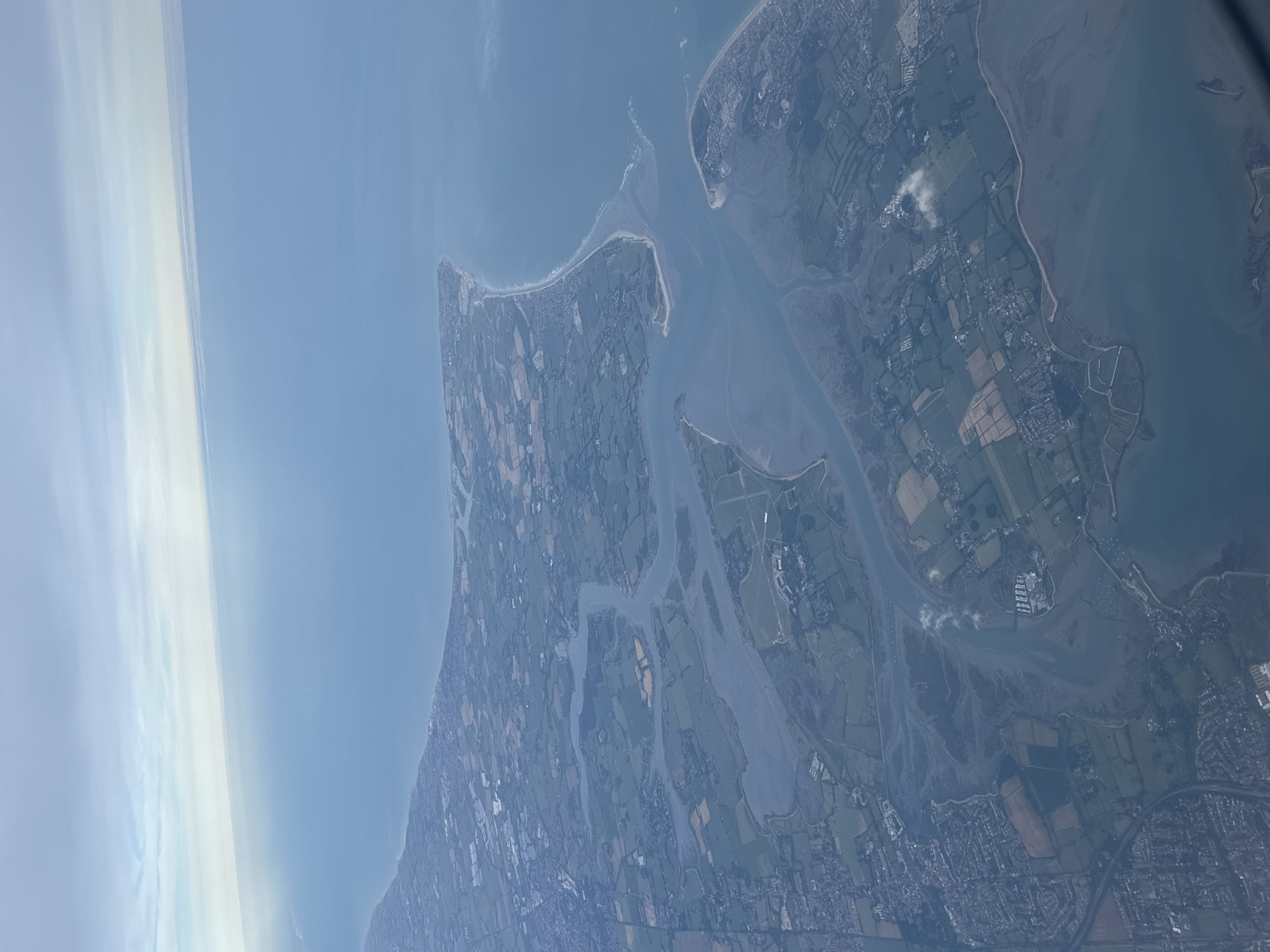 An aerial image captures a detailed view of the coastline and estuarine system around Wexford, County Wexford, Ireland. The scene unfolds under a pale blue sky with faint, wispy cloud formations.
The upper and right portions of the image are dominated by the open sea, the Irish Sea, appearing as a vast expanse of blue-grey water. The coastline curves sharply inwards, revealing a complex estuarine environment.
At the center, Wexford Harbour is prominently featured, a large, partially enclosed body of water fed by the River Slaney and other waterways. The water within the harbour appears turbid and silt-laden, with extensive mudflats and shallow areas visible, particularly along its western and southern boundaries.
The entrance to Wexford Harbour, located towards the right, is formed by two distinct landforms:
1. **Northern side (top right of harbour entrance):** Raven Point Nature Reserve and Curracloe Beach, appearing as a long, curving sandy spit of land, densely vegetated with trees, extending into the sea. A long, light-colored sandy beach is visible along its outer edge. Scattered development is present along this coastal stretch.
2. **Southern side (bottom right of harbour entrance):** Rosslare Strand, another elongated sandy spit, with residential and commercial development (the town of Rosslare) situated further south along the coast.
To the left and bottom-left, the land comprises a mosaic of rural and urban landscapes. Large areas are dedicated to agriculture, visible as geometrically shaped fields in varying shades of green and brown, indicating cultivation. Interspersed within this rural fabric are dense urban and suburban areas, notably Wexford town in the lower-left quadrant. Here, a grid of roads and numerous residential and commercial buildings are discernible. Further inland, multiple rivers and tidal channels branch off the main harbour, winding through the cultivated fields and creating smaller inlets and marshy areas.
No individual people or specific actions are discernible due to the high altitude of the photograph. The scene emphasizes the intricate interaction between natural coastal processes and human development in a temperate coastal zone.