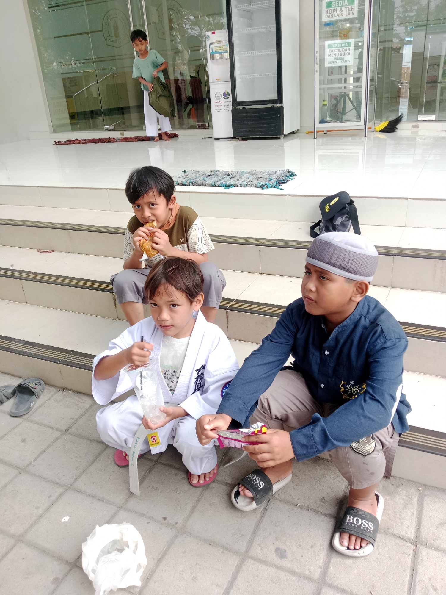 Three boys are sitting on concrete stairs in front of a building in Kediri City, Indonesia. The boy on the left is wearing a white karate gi and holding a drink with a straw. The boy in the middle is wearing a brown and white patterned shirt and grey pants, and he is eating something. The boy on the right is wearing a blue long-sleeved shirt, grey pants, and a grey and white cap. He is looking to the right and holding a candy bar. A black backpack is visible on the stairs behind the boy in the middle. In the background, there is a glass-fronted building with signs, a water dispenser, and a large refrigerator.