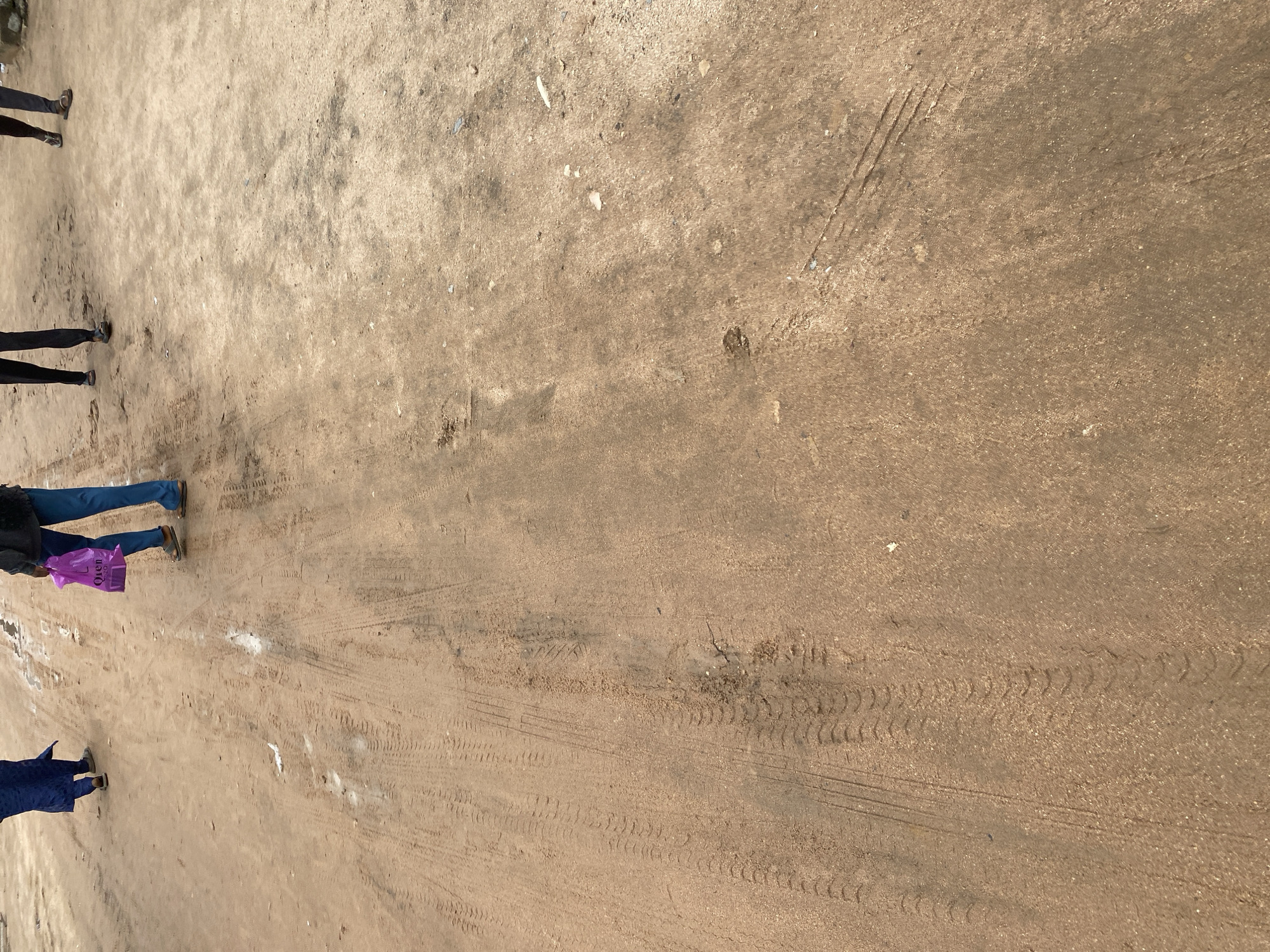 This media file captures an overhead view of a sandy or dirt ground with several individuals standing or walking on it. The ground displays numerous tire tracks, indicating vehicular activity.
The subjects visible are the lower halves of at least four people, dressed in casual clothing. One person on the left is wearing dark pants and appears to be wearing sandals. Another person, closer to the center and slightly below, is wearing blue jeans and is carrying a bright purple shopping bag. A third person, further down and to the left, is wearing a dark blue garment and sandals. The fourth person, at the very top left, is only partially visible, wearing dark pants and sandals.
The scene appears to be outdoors in a location that could be a road, a path, or an open area with loose ground. The presence of tire tracks suggests it's a place frequented by vehicles. The lighting indicates it is daytime, likely under clear or partly cloudy skies, as there are no strong shadows suggesting a particular time of day.
The overall mood is neutral, focusing on the mundane activity of people on a dusty surface. No specific event is clearly depicted, but it seems to be a moment in time where individuals are traversing this area. There is some visible white debris scattered on the ground in patches. The text "Olen" is visible on the purple shopping bag. Given the provided location context of Jalingo, Nigeria, the scene is likely taking place within that region.