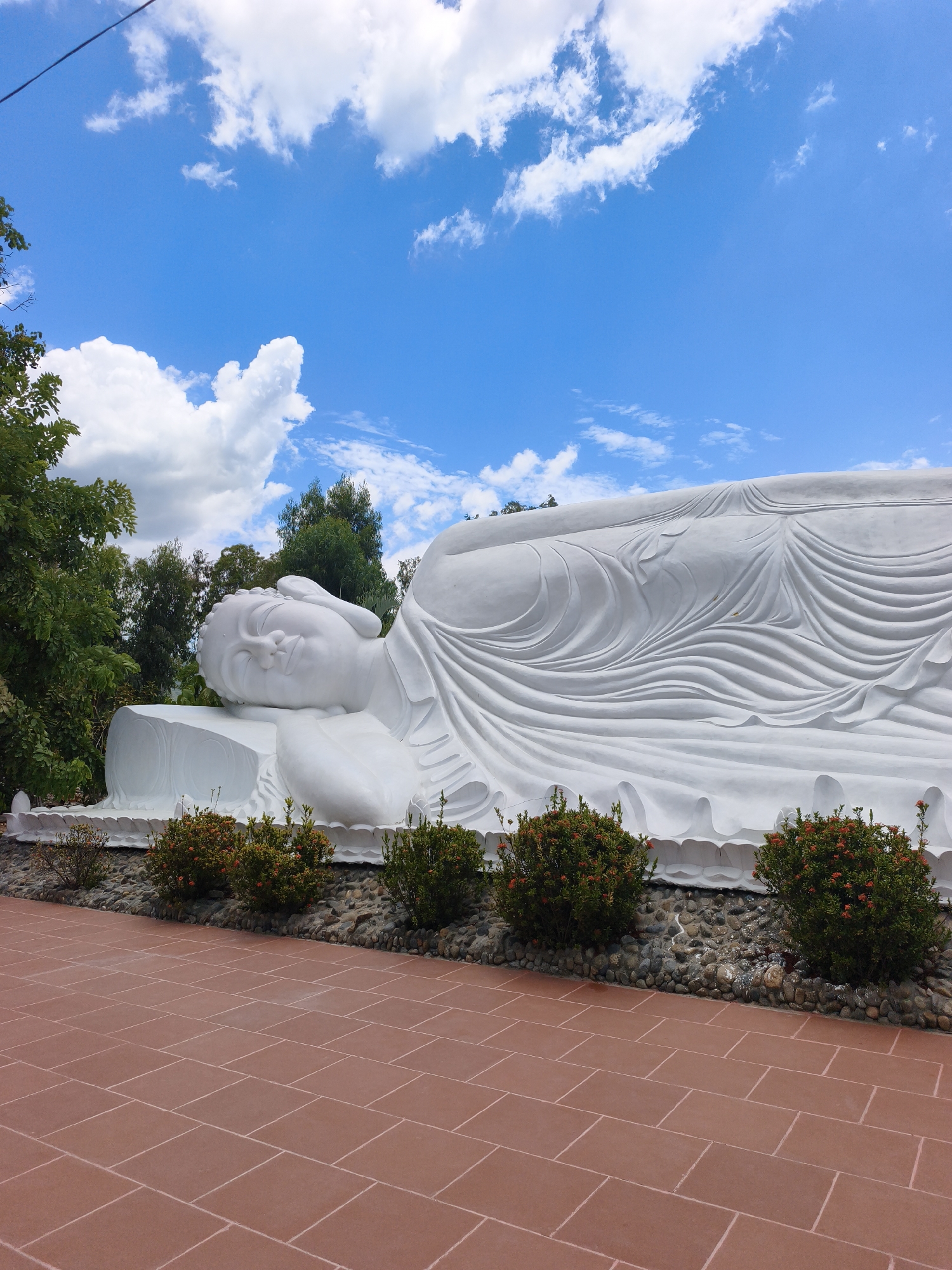 A monumental white reclining Buddha statue is the central subject of this image. The statue is positioned outdoors, with lush green trees and a vibrant blue sky dotted with fluffy white clouds in the background. The foreground features a reddish-brown tiled pathway, a decorative border of small stones, and several low-lying green bushes with small red flowers. The statue itself is detailed, with folds in its garment artfully sculpted and a serene expression on its face. This location is the Quan Am Tu Thi Buddhist temple in Cam Ranh, Vietnam, known for its impressive reclining Buddha.