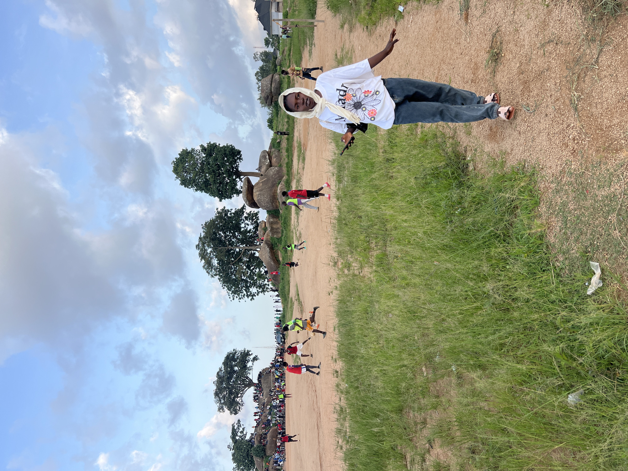 The image depicts a lively outdoor scene in Jos, Nigeria, likely on a warm afternoon, given the cloud cover. In the foreground, a young woman walks towards the camera, holding a smartphone in her right hand. She is wearing a white t-shirt with a floral design and the word "Madr" printed on it, dark blue jeans, and a light-colored headscarf. Her left hand is extended outwards as she walks along a dirt path.
The background reveals a busy football (soccer) match in progress on a sandy field. Numerous people are gathered, some watching the game from the sidelines, while others are actively playing. Spectators are positioned on grassy hills and rocky outcrops, suggesting a natural amphitheater-like setting. Several trees, some with dense foliage and others more sparse, are scattered throughout the landscape, with large rock formations visible in the middle ground and background, characteristic of Jos's topography. The sky is filled with a mix of blue sky and fluffy clouds, hinting at a partly cloudy day. A few buildings and what appears to be a volleyball net are visible on the right side of the frame, indicating the presence of a community or recreational area. The overall impression is one of a vibrant community event taking place in a scenic, natural environment.