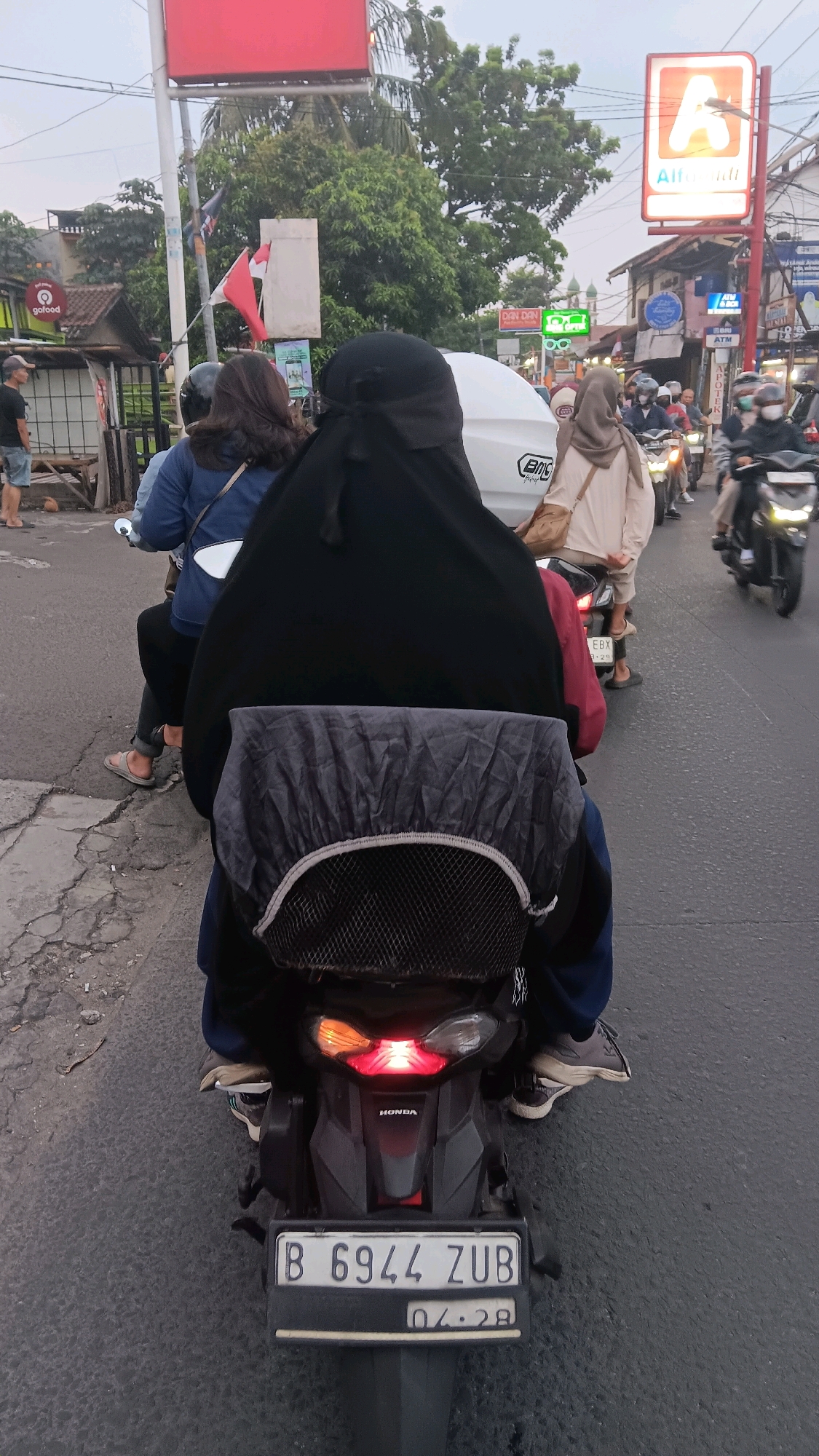 An overhead shot captures a motorcycle traffic scene on a street in Depok, Indonesia, likely on a cloudy day. The focus is on a motorcycle carrying two individuals. The rear passenger, wearing a black niqab and headscarf, is covered by a dark grey rain cover over a black backpack. The driver, in a blue jacket, is partially visible. The motorcycle's license plate reads "B 6944 ZUB" and shows an expiration date of "04.28". Other motorcycles and pedestrians are present in the background, with signs of commercial establishments visible, including an "A" logo for Alfamart. The road surface is asphalt, with some cracks and debris. The environment suggests an urban setting with moderate traffic.