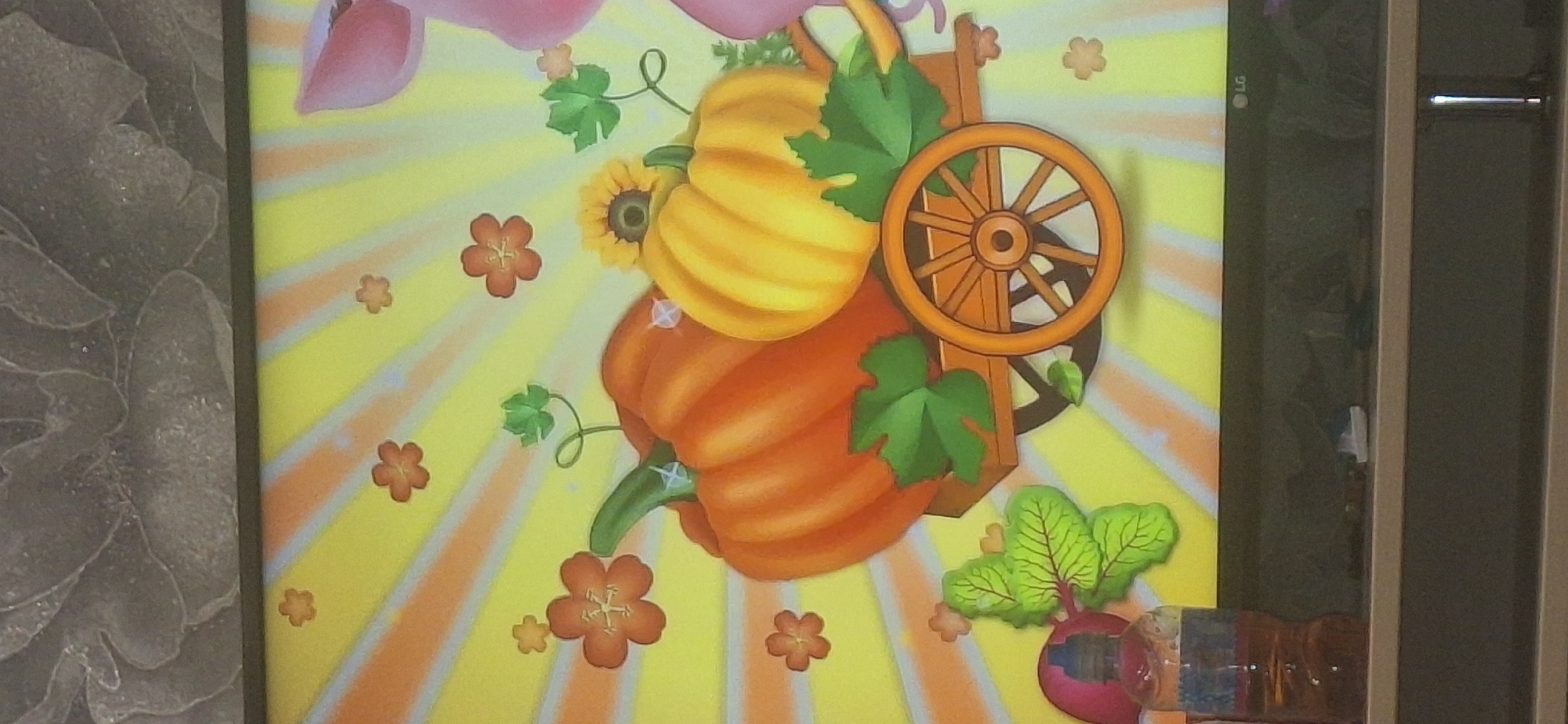 The image shows a close-up of a television screen displaying a cartoonish illustration. The illustration features two pumpkins, one large orange and one smaller yellow, on a wooden cart with a spoked wheel. There are also several small, stylized flowers and green leaves scattered around the pumpkins. The background of the illustration is a bright yellow with radiating orange and white lines, suggesting a sunny day. In the lower left corner of the screen, a plastic bottle containing a brown liquid is partially visible. The bottom of the television screen shows the LG logo. The overall scene is brightly colored and appears to be aimed at a young audience. The image was taken indoors, likely in a residential setting, and there is no discernible connection to Bila Tserkva, Ukraine.