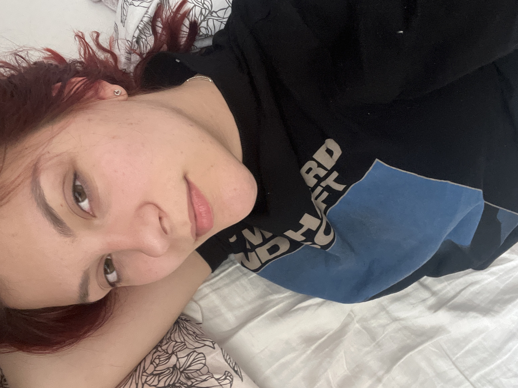 A young, fair-skinned woman with reddish-brown hair lies on her side on a bed, facing the camera. She wears a dark sweatshirt with a visible blue and white design. The bed has white sheets and a patterned pillow. The location cannot be determined from the image.